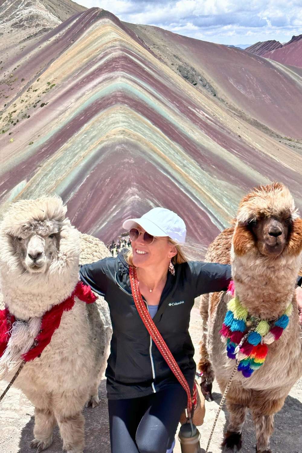 This image captures Kate smiling between two alpacas, both adorned with colorful decorations, with the vibrant stripes of Rainbow Mountain (Vinicunca) in Peru as the stunning backdrop.