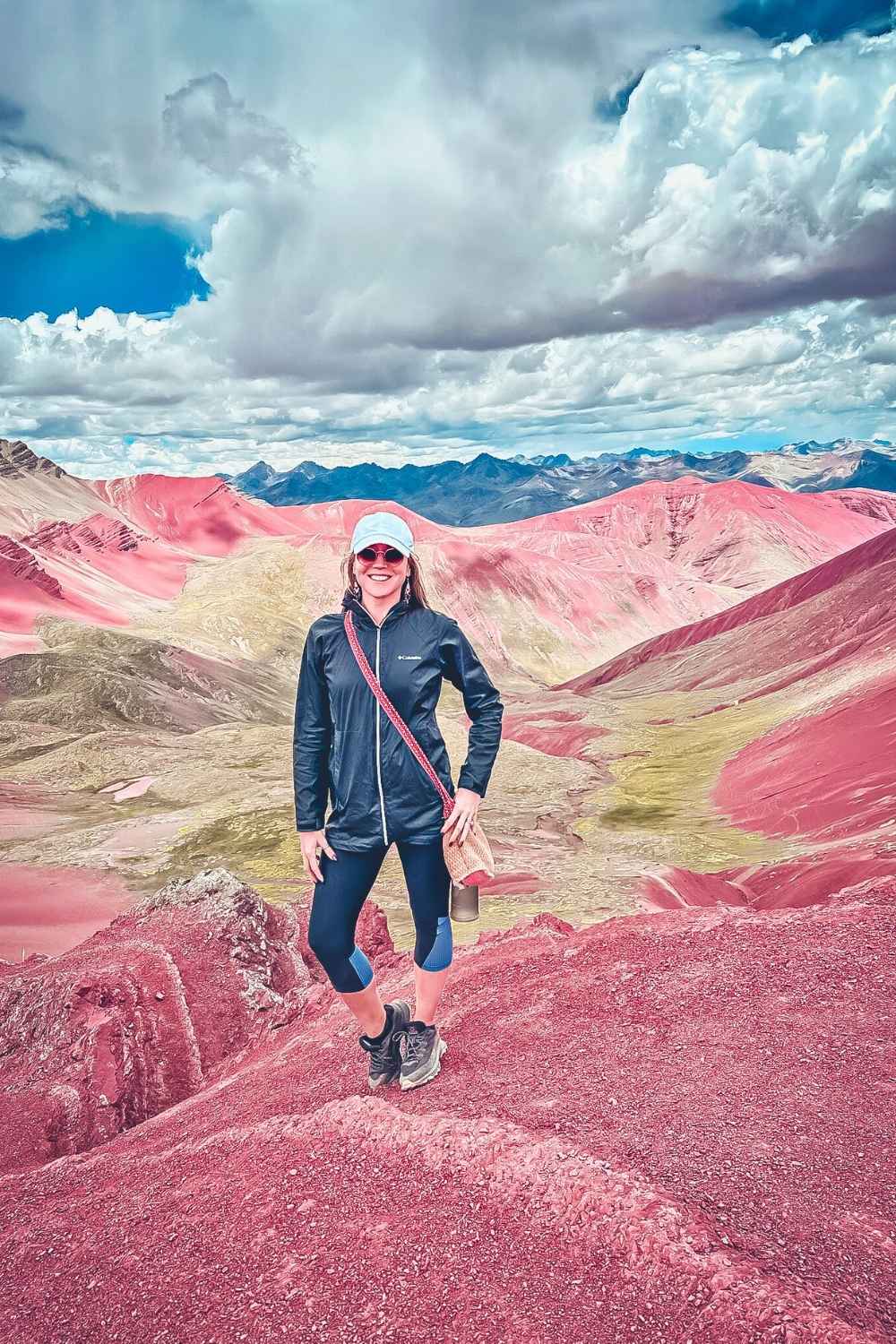 This photo shows Kate standing confidently in the vibrant Red Valley (Valle Rojo) in Peru, surrounded by striking red and ochre-colored hills under a dramatic sky filled with fluffy clouds. The unique landscape highlights the raw, untouched beauty of this high-altitude Andean region.