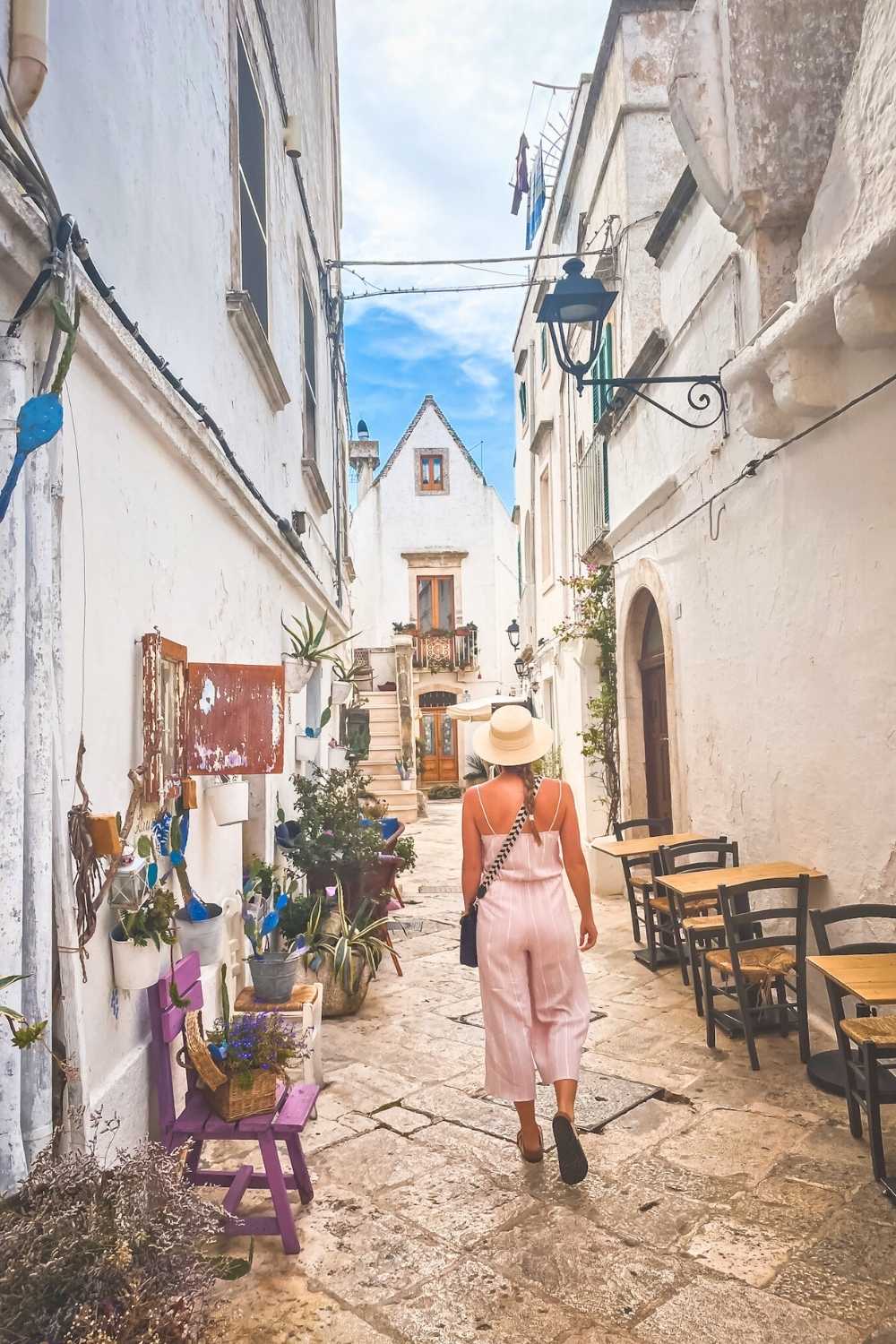 This photo captures Kate strolling down a charming cobblestone alley in Puglia, Italy, surrounded by whitewashed buildings adorned with colorful potted plants and rustic decor. The tranquil scene highlights the region's Mediterranean charm.