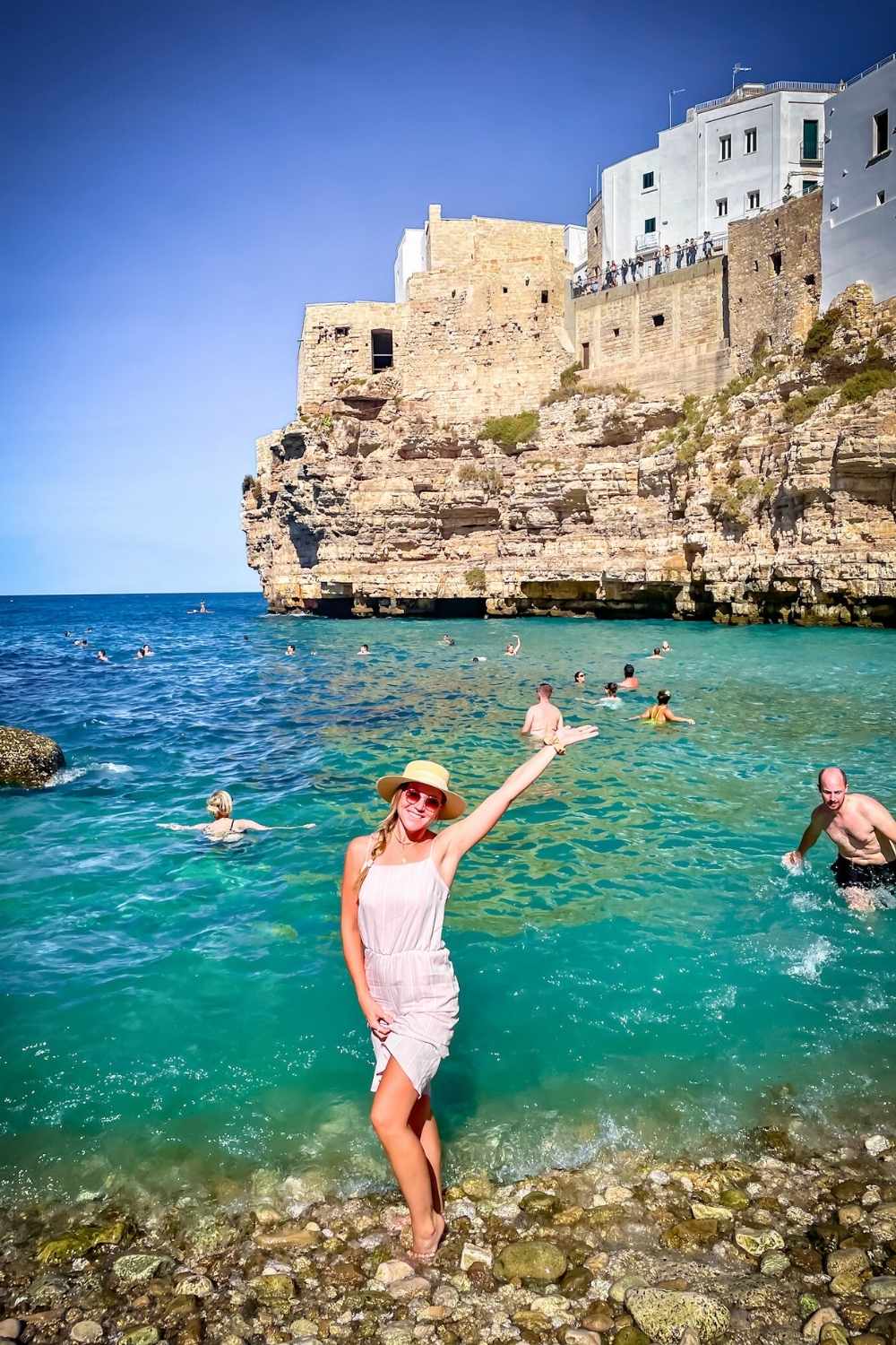 This photo features Kate enjoying the crystal-clear waters of a rocky beach in Puglia, Italy, with the backdrop of historic stone buildings perched on cliffs. The vibrant turquoise sea and the sunlit architecture showcase the stunning coastal charm of the region.