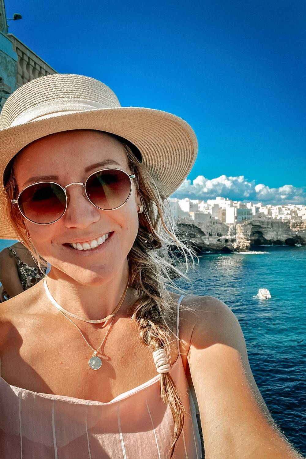 This close-up photo captures Kate wearing sunglasses and a straw hat, with their hair in a braid. In the background, the bright blue sea and white stone buildings perched on cliffs beautifully showcase the coastal charm of Puglia, Italy, under a vibrant sky.