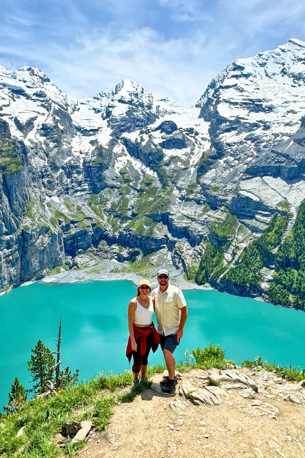 This vibrant photo captures Kate and her husband standing on a scenic overlook with the turquoise waters of Oeschinensee Lake below, surrounded by dramatic, snow-capped Swiss Alps. The lush greenery and rugged cliffs create a striking contrast, showcasing the natural beauty of Switzerland.