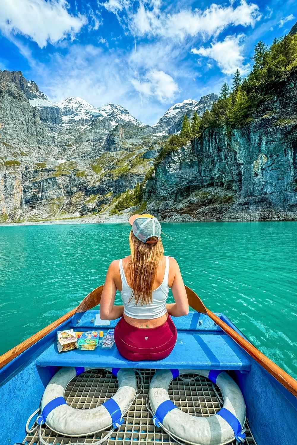 This vibrant image showcases Kate seated in a small boat on the crystal-clear turquoise waters of Oeschinensee Lake, Switzerland. She faces a stunning backdrop of towering cliffs, snow-capped mountains, and lush greenery under a bright blue sky. Snacks are visible on the bench in front of her.