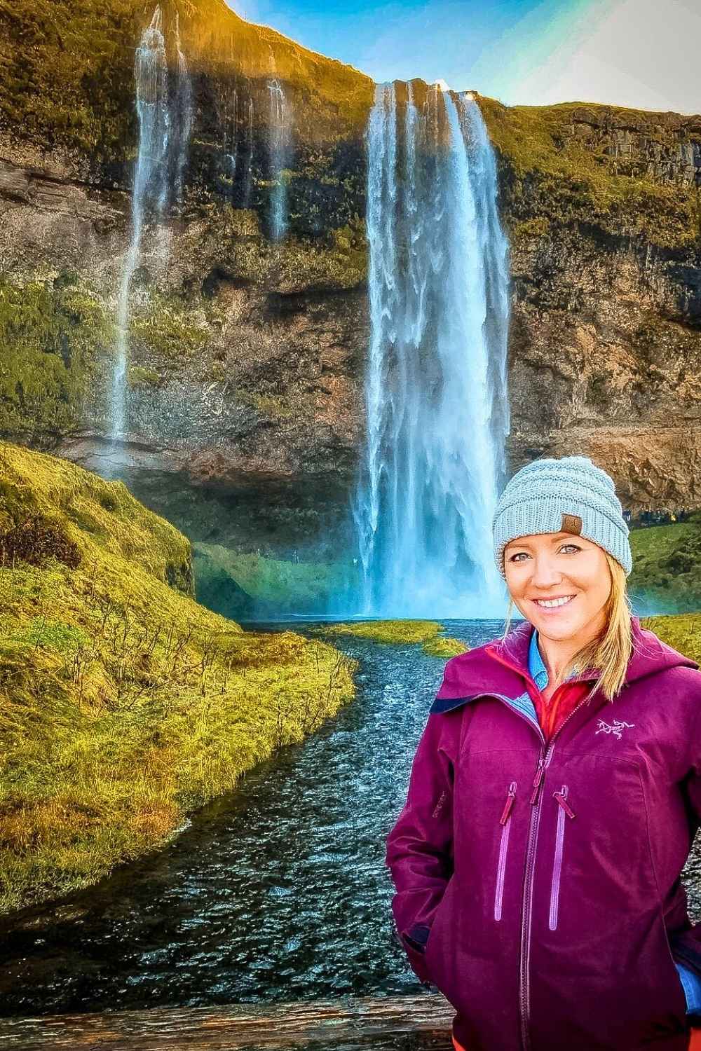 This image showcases Kate in a purple jacket and blue beanie standing in front of the stunning Seljalandsfoss waterfall in Iceland. The cascading water flows gracefully down moss-covered cliffs, with sunlight adding a golden hue to the scene. 