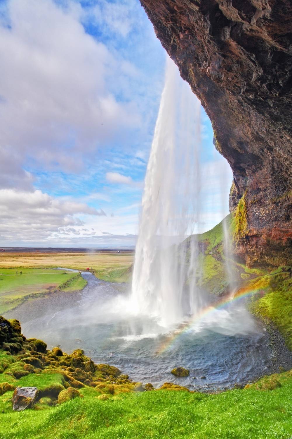 This photo features the iconic Seljalandsfoss waterfall in Iceland, captured from behind the cascading curtain of water. The vibrant green moss and grass-covered cliffs add a lush contrast to the roaring falls, while a faint rainbow arches over the spray.