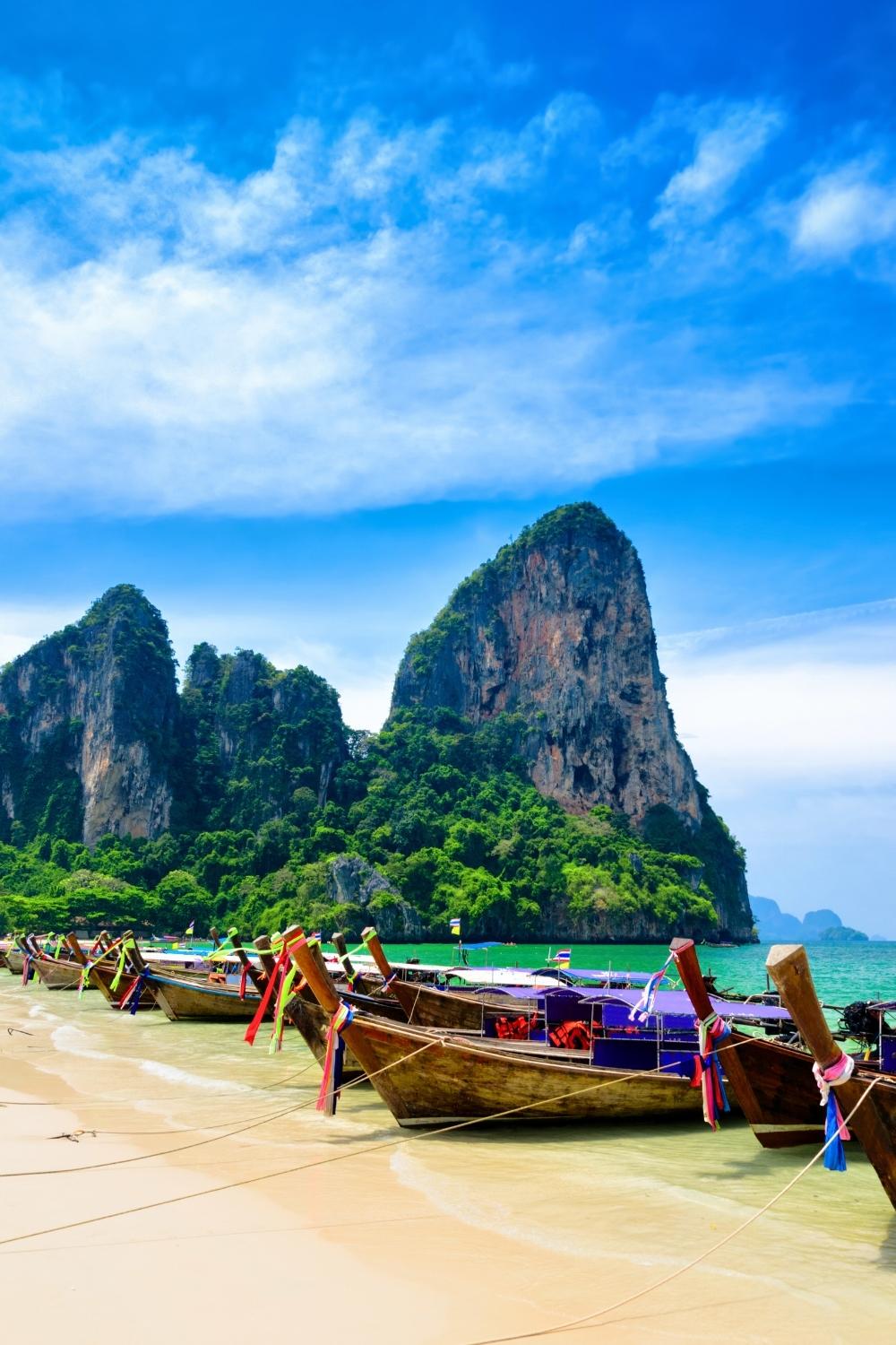 This image showcases traditional Thai longtail boats lined up along a pristine sandy beach with turquoise waters. Towering limestone cliffs covered in lush greenery dominate the background, under a bright blue sky with wispy clouds, evoking the serene beauty of southern Thailand's coastal landscape.