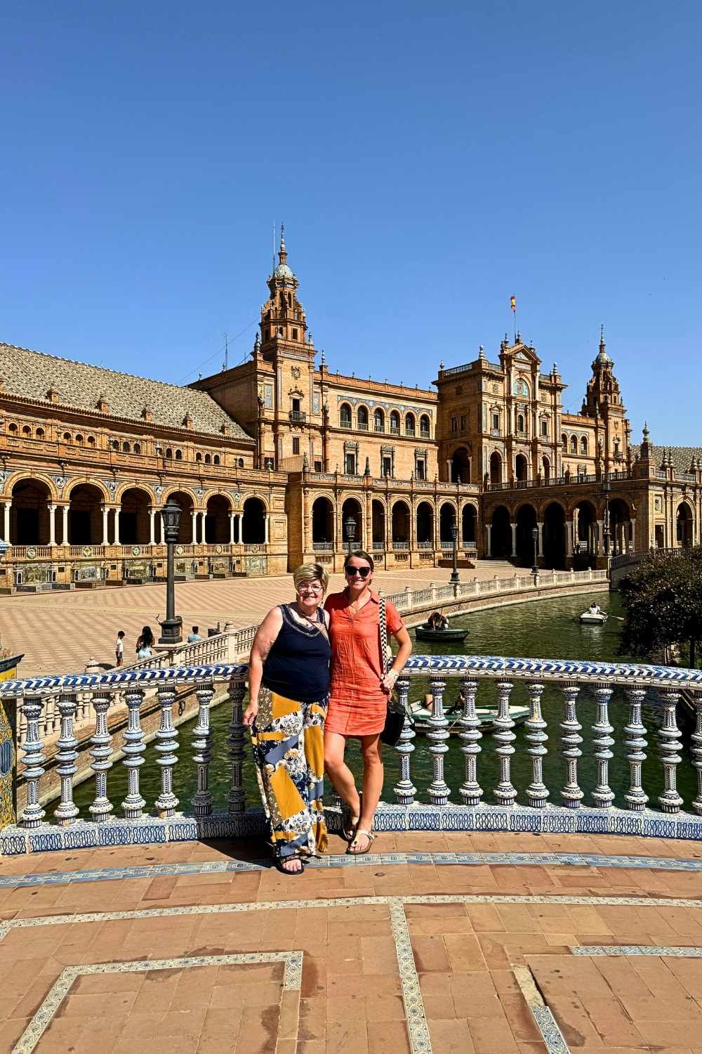 This photo shows Kate and her mother posing on a beautifully tiled bridge at Plaza de España in Seville, Spain. The grand semi-circular building with its intricate architecture, arched colonnades, and central canal creates a stunning backdrop under a clear blue sky.