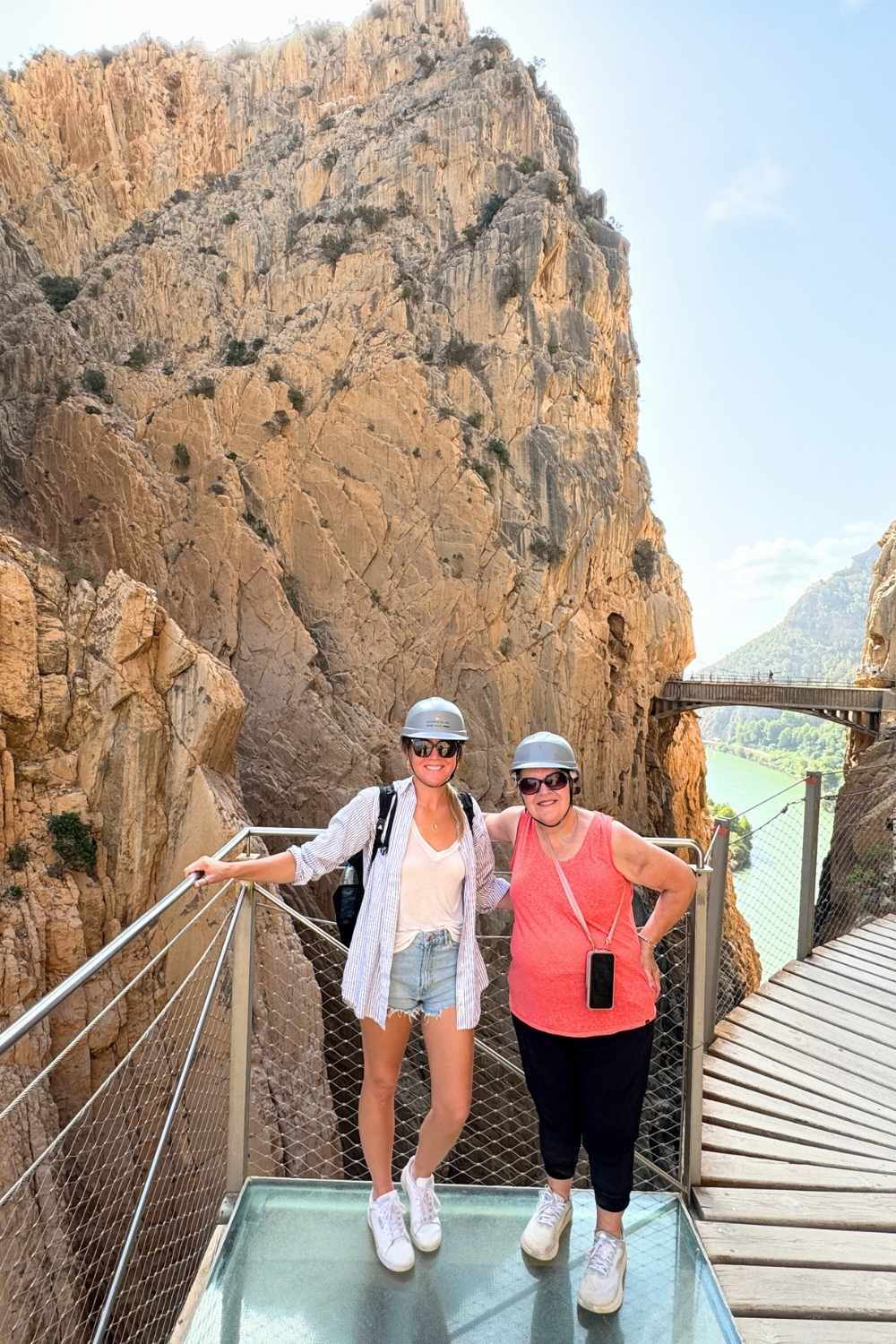 This photo shows Kate and her mother standing on a glass-bottom platform along the Caminito del Rey hiking trail in Spain. They are both wearing helmets and smiling against a backdrop of towering rocky cliffs and a wooden walkway overlooking a turquoise river below.