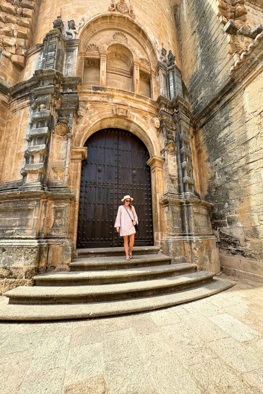 This photo captures Kate standing in front of an ornate, historic wooden door framed by detailed stonework in Andalucía, Spain. She is wearing a light dress, a sun hat, and sunglasses, stepping gracefully down the curved stone steps of the entrance.