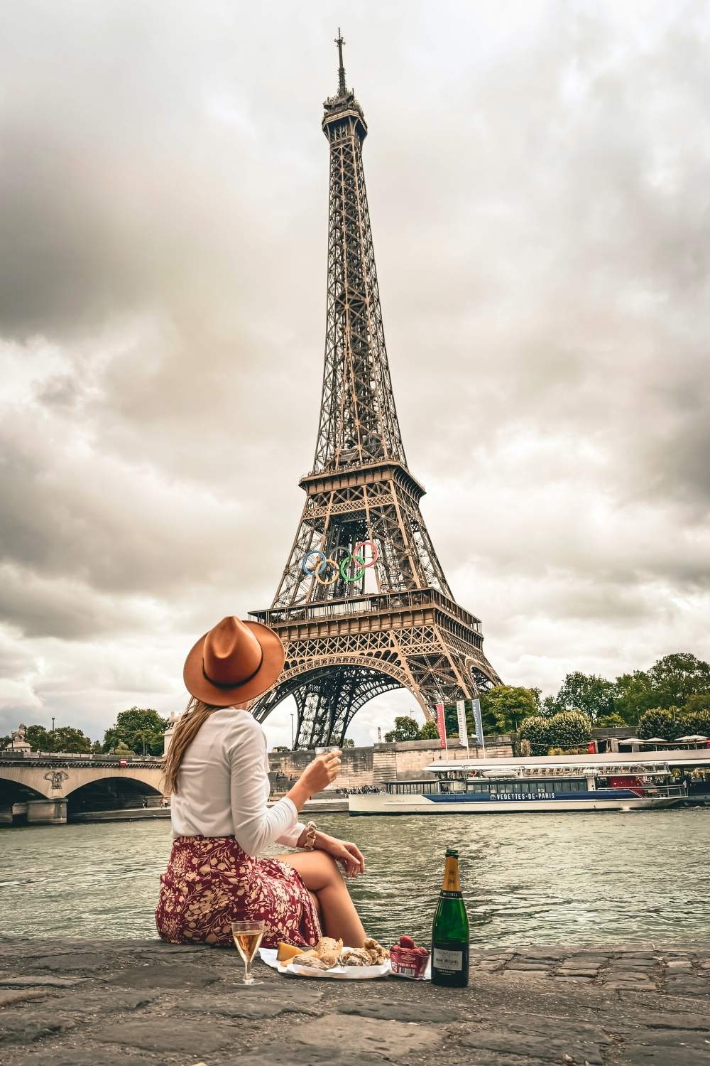 This picturesque image shows Kate sitting along the Seine River in Paris, with the Eiffel Tower prominently in the background under a cloudy sky. She is enjoying a picnic spread of champagne and treats, wearing a wide-brimmed hat, a white top, and a patterned skirt, embodying the Parisian charm.