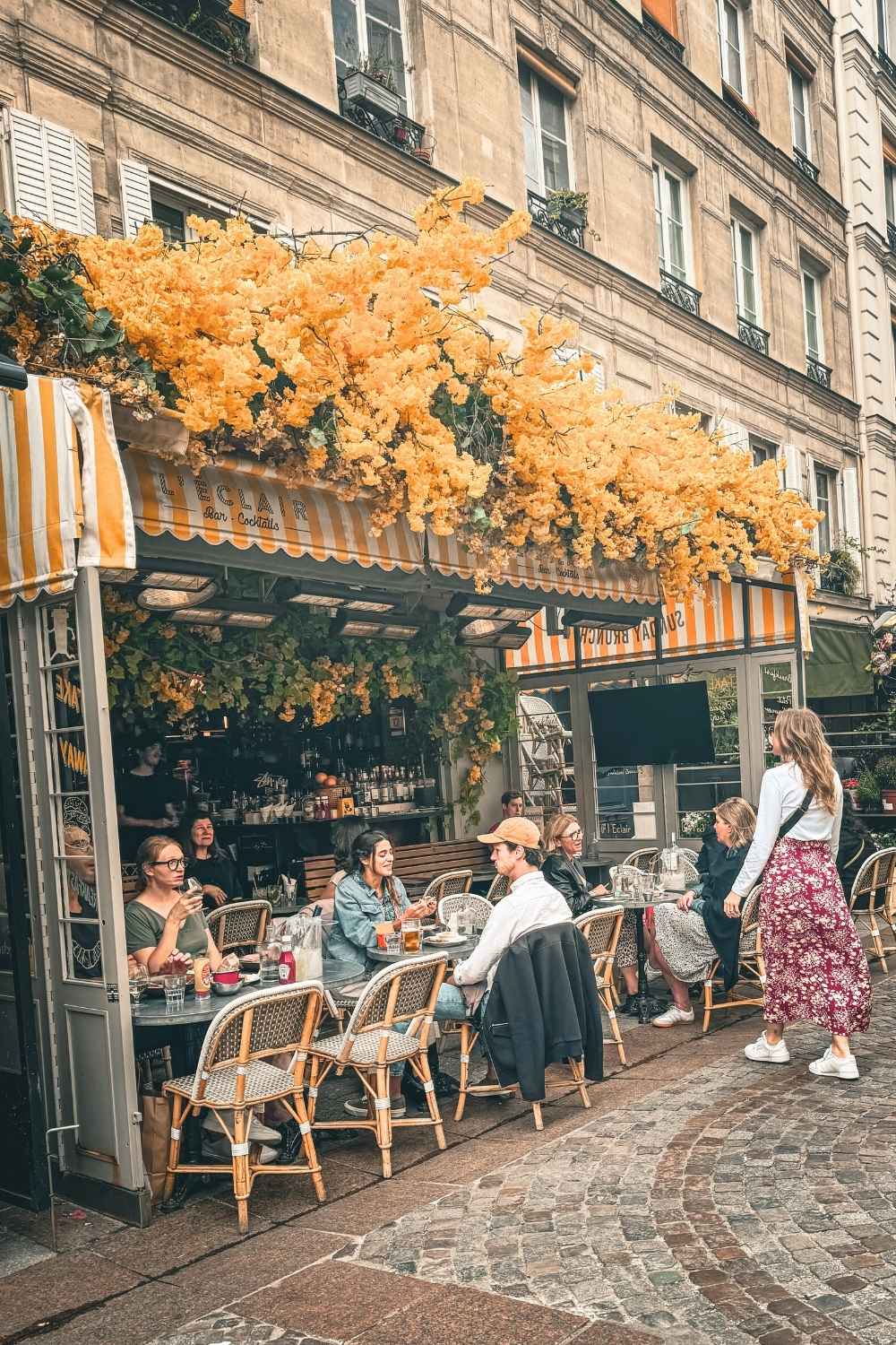 This vibrant photo captures Kate in a lively Parisian café scene, surrounded by patrons enjoying their meals under an awning adorned with cascading yellow flowers. Kate, in a white top and floral skirt, blends seamlessly into the charming and bustling ambiance of the cobblestone street.