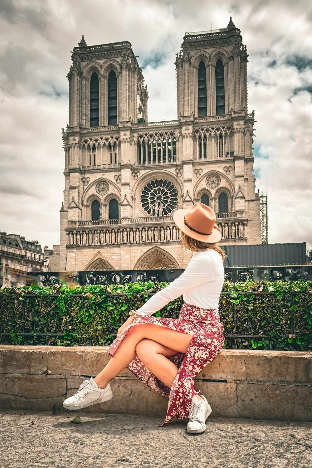 This photo features Kate sitting gracefully in front of the iconic Notre-Dame Cathedral in Paris. Wearing a stylish white top, a floral skirt, and a brown hat, she gazes up at the majestic Gothic architecture, adding a serene and fashionable touch to the historic backdrop.