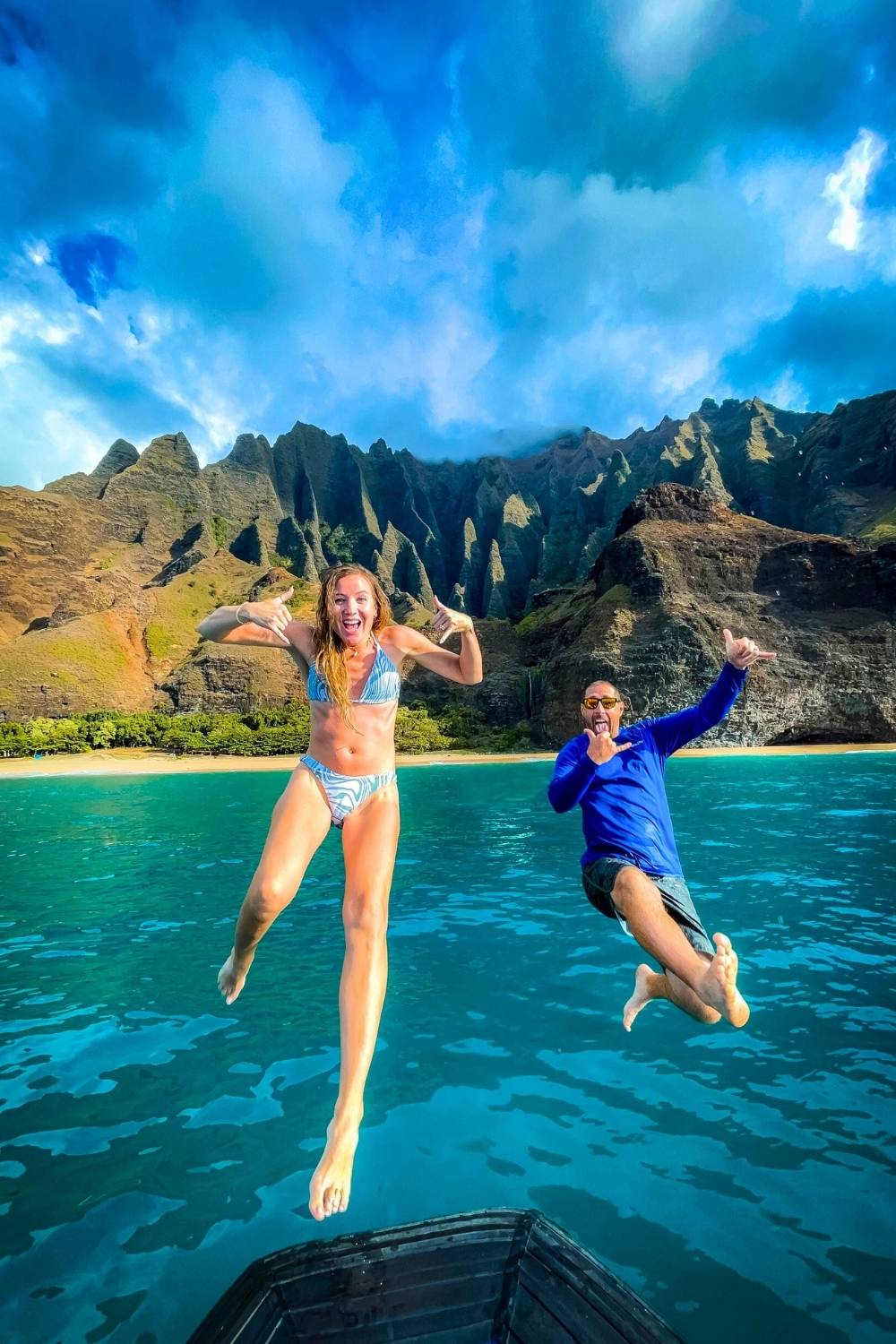 The image captures Kate and her husband mid-jump off a boat into the clear turquoise waters of the Na Pali Coast in Kauai, Hawaii. The dramatic backdrop showcases the rugged green cliffs and a secluded beach under a bright blue sky.