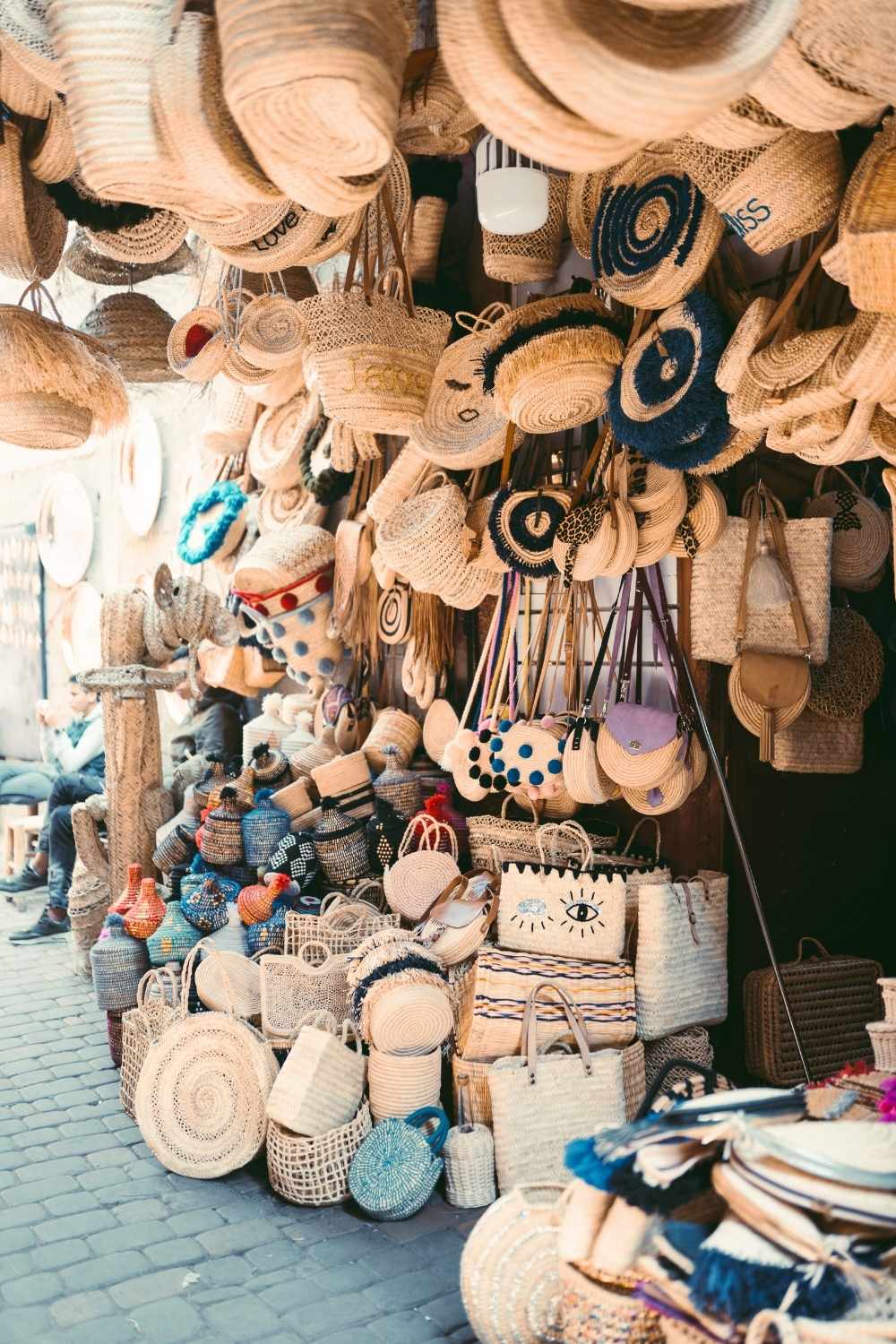 This image captures a vibrant market stall in Marrakesh, Morocco, showcasing a variety of handwoven baskets, hats, and bags made from natural materials. The intricate designs and bright embellishments reflect the rich artisan culture of the region, with sunlight streaming in to highlight the textures. 