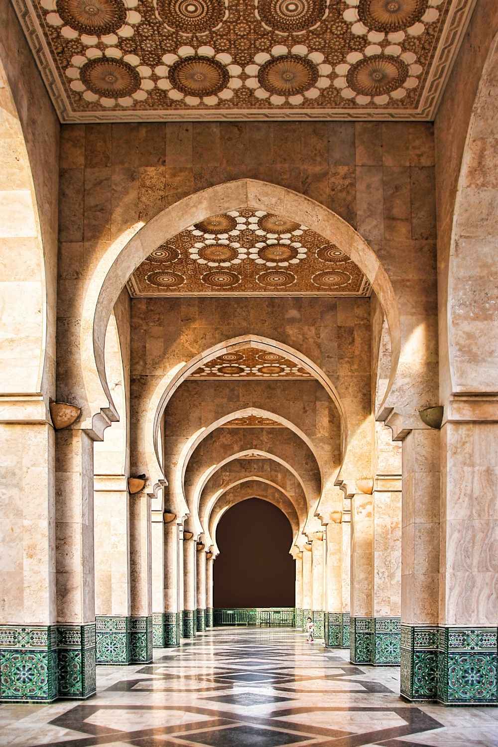 This image features the stunning architecture of Marrakesh, Morocco, showcasing intricately carved arches and geometric patterns on the ceiling. The marble floors reflect the sunlight streaming through the columns, creating a serene and symmetrical scene. This space highlights traditional Moroccan craftsmanship and architectural elegance.