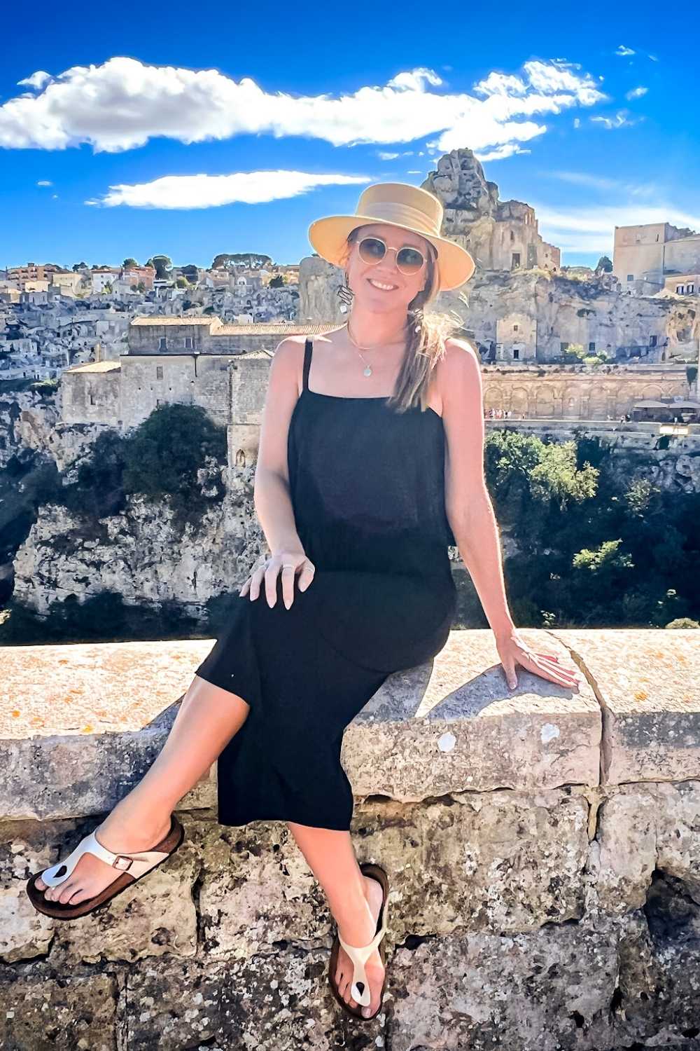 This photo captures Kate sitting on a stone ledge overlooking the ancient rock city of Sassi di Matera in Italy. She is dressed in a black outfit, wearing a straw hat and sunglasses, with the historic cliffside buildings and bright blue sky creating a stunning backdrop.