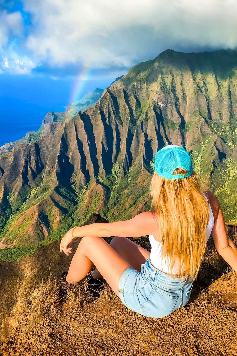 This image features a scenic view of the Na Pali Coast in Kauai, Hawaii, with its dramatic cliffs and lush green ridges extending toward the blue ocean. Kate is wearing a blue cap, white tank top, and denim shorts sits on a rocky ledge, gazing at the landscape with a faint rainbow in the distance. The vibrant colors and natural beauty emphasize the majesty of this iconic Hawaiian location.