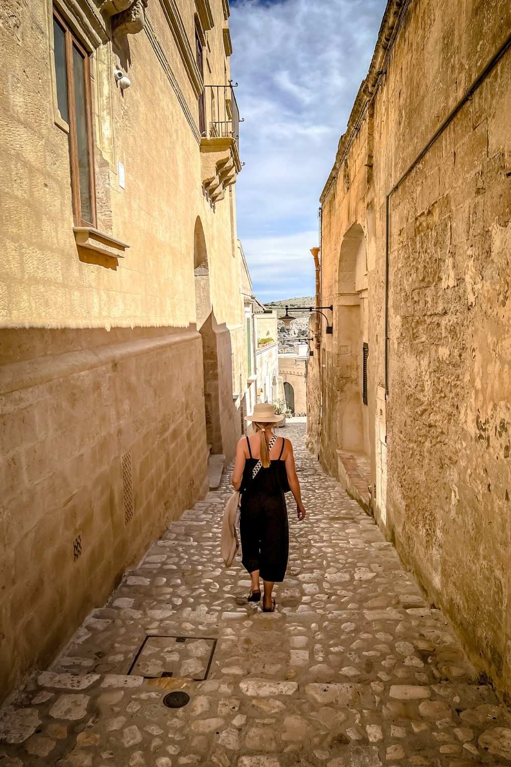 This image shows Kate walking down a narrow cobblestone alley in Matera, Italy. She is wearing a black jumper with a staw hat.