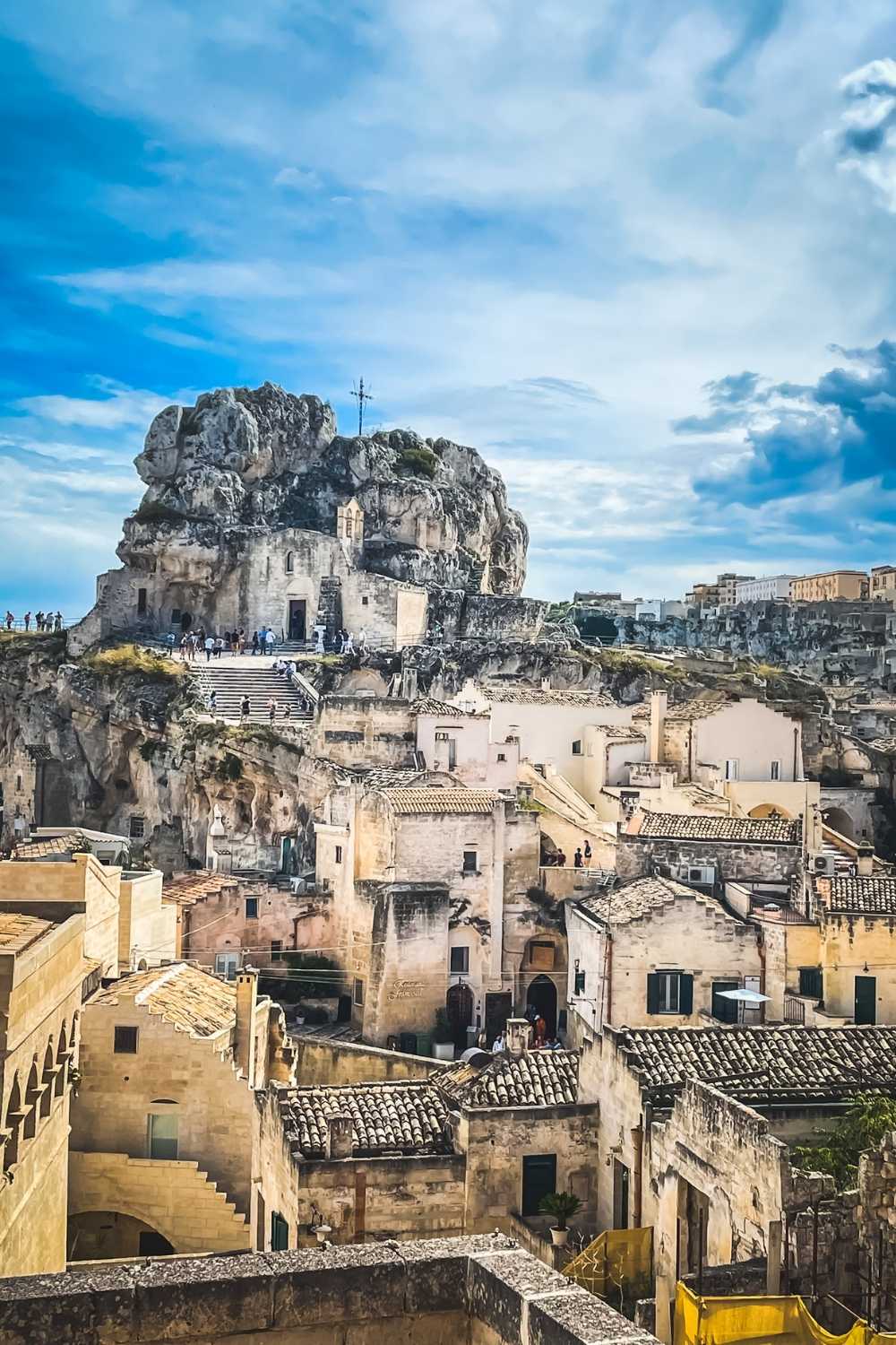 This photo showcases the historic architecture of Matera, Italy, with its iconic cave dwellings and stone buildings nestled into the rocky landscape. The centerpiece is a large rock formation topped with a small church, surrounded by terracotta roofs and stone pathways.