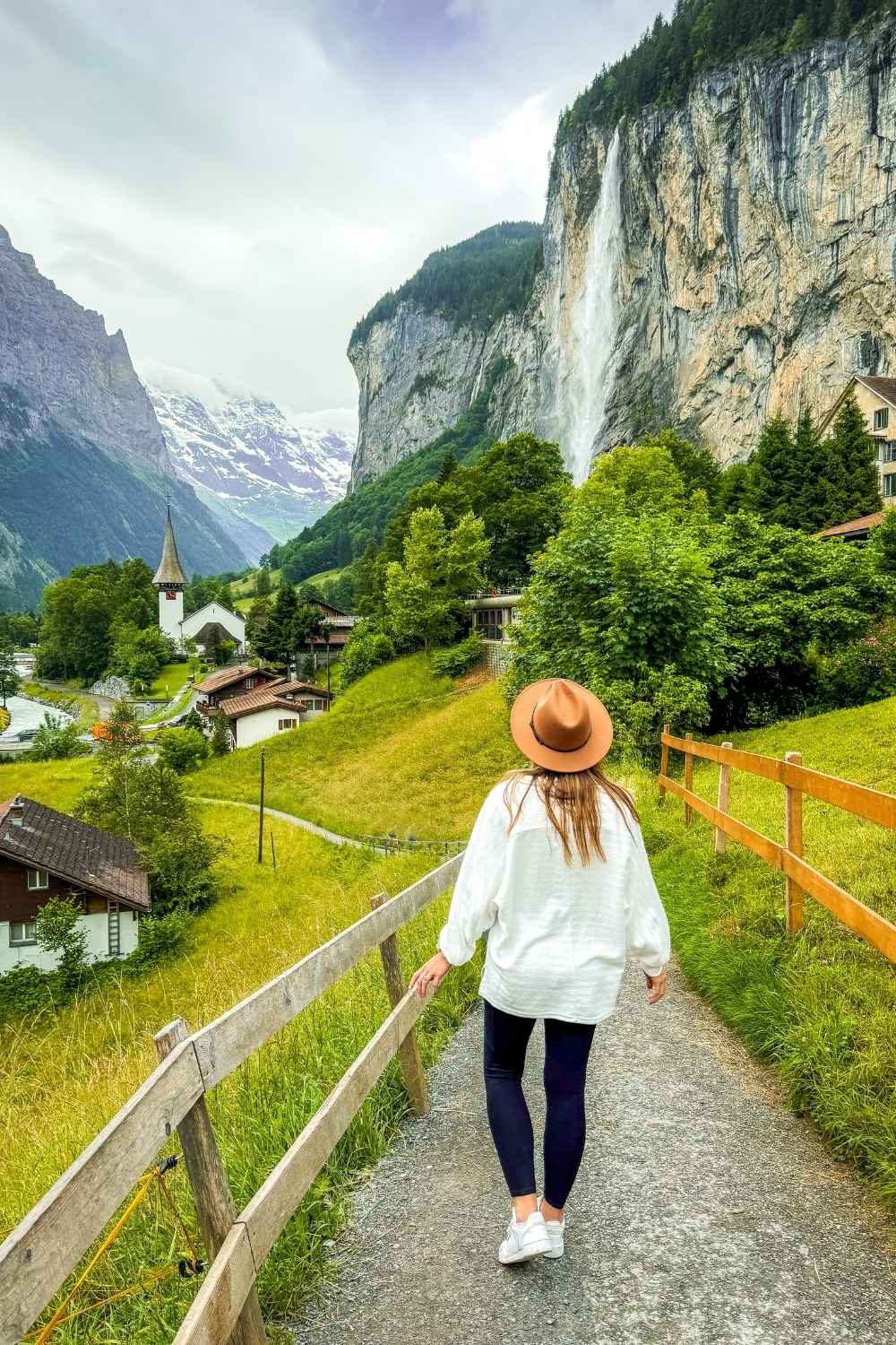 This photo captures Kate walking down a narrow walkway leading towards the towering cliff side waterfall in the center of Lauterbrunnen, Switzerland. She is wearing a white long sleeve sweater, brown hat and black pants. The landscape is lush with greenery and it appears to be drizzling rain. 