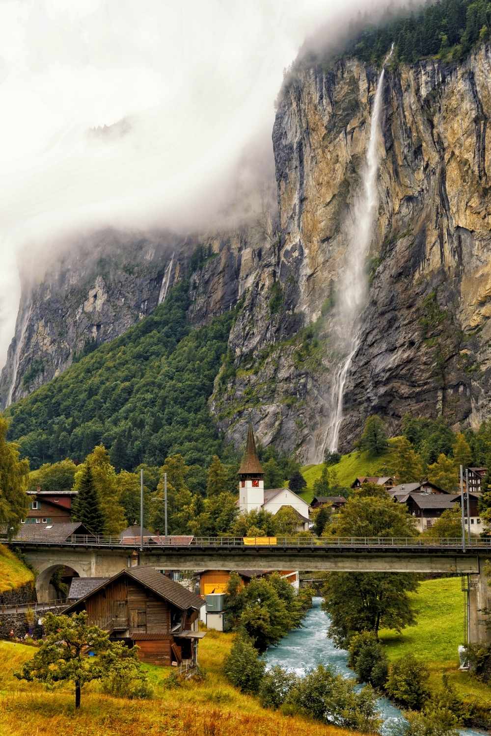 This photo showcases the lush Lauterbrunnen town surrounded by towering cliffs, lush greenery and a dramatic waterfall flowing in the middle of the town. There is a river rushing though the center of town with a white stapled church in the background. 