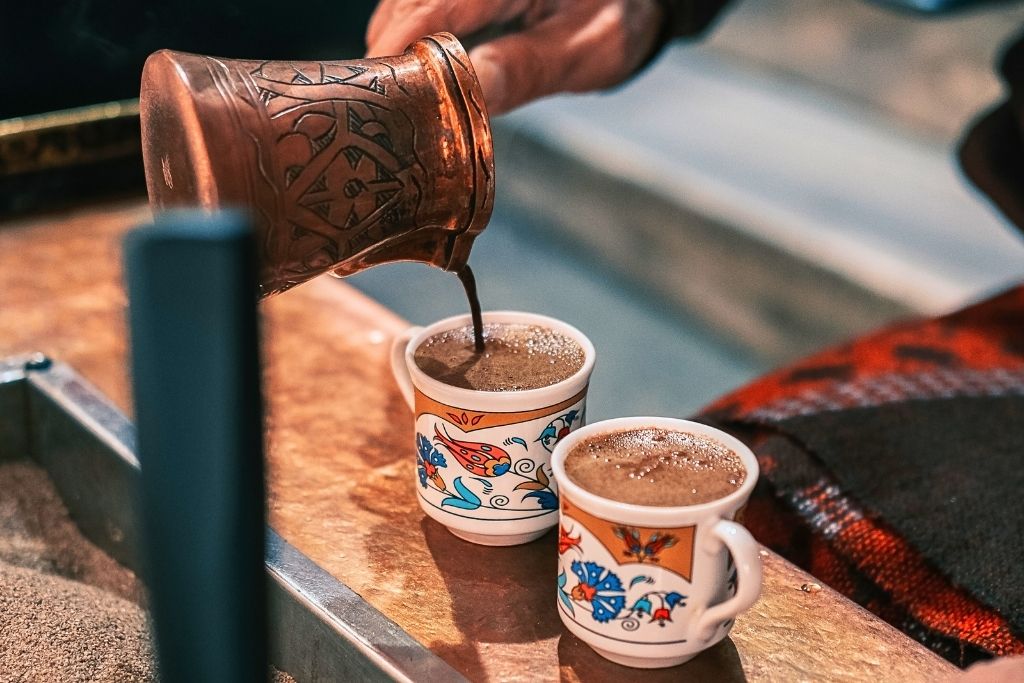 This image shows two small, ornately decorated cups filled with frothy Turkish coffee, with coffee being poured from a traditional copper cezve. The setting evokes the cultural charm of a food tour in Istanbul, showcasing the rich coffee tradition.