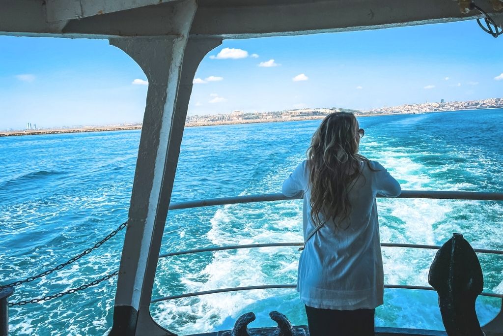 This image captures a serene moment Kate gazes out over the Bosphorus Strait from the deck of a ferry. The deep blue water reflects the bright sky, with the cityscape of Istanbul visible in the distance. The scene highlights the beauty and tranquility of crossing the iconic waterway that divides Europe and Asia.