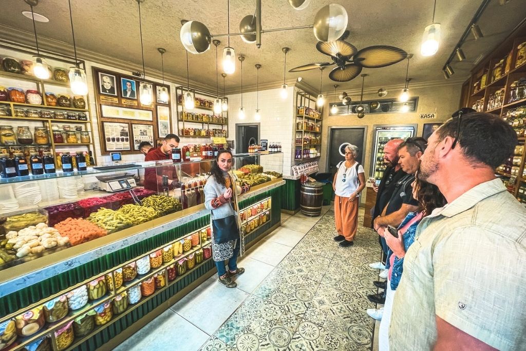 This image captures the interior of a colorful pickled goods shop during a food tour in Istanbul, with jars of pickles and preserved foods neatly displayed on shelves and counters. A guide appears to be explaining something to a group of people who are attentively listening, adding to the immersive culinary experience. The shop’s vibrant decor and wide selection highlight Istanbul’s rich food culture.