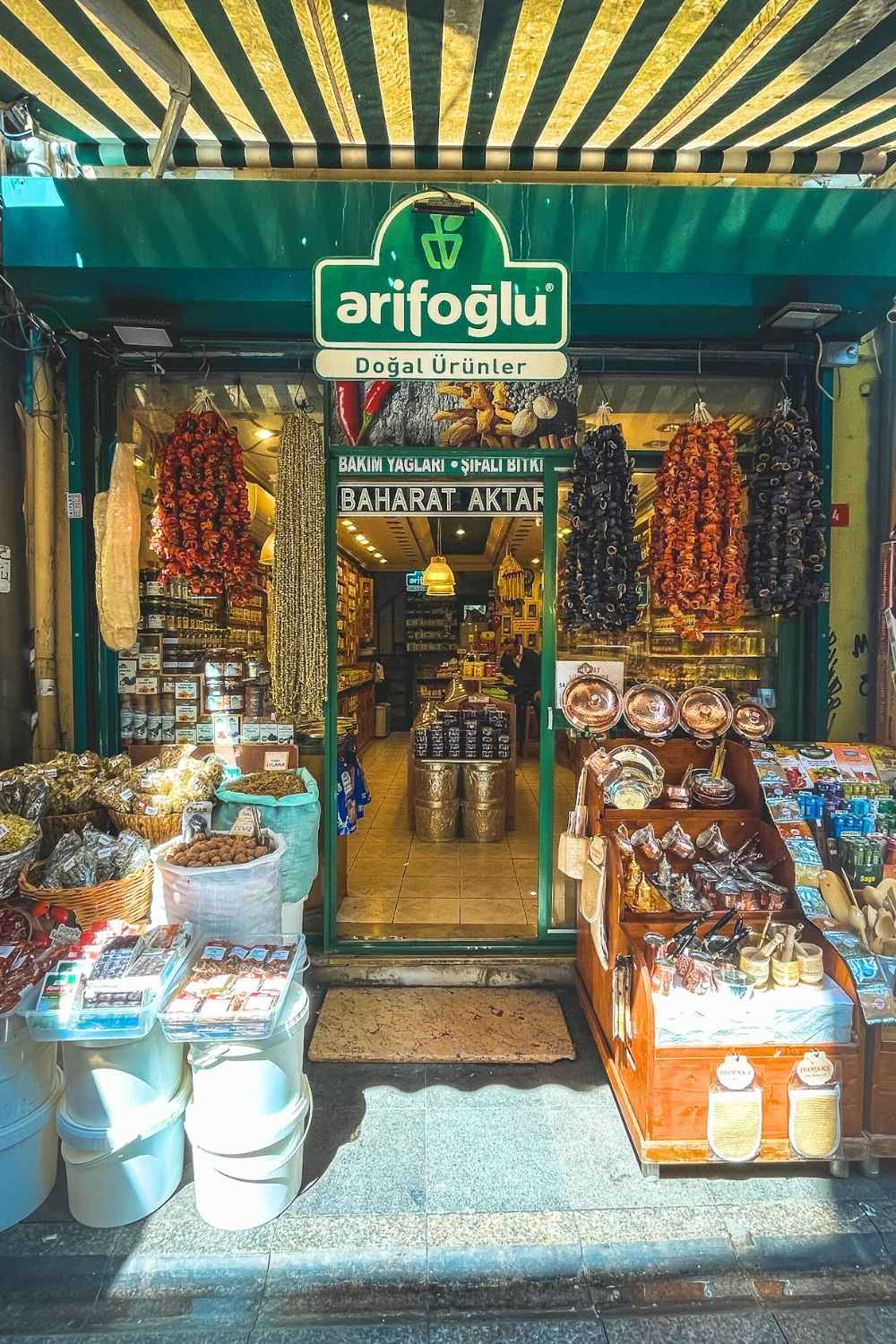 This image shows the entrance of a spice and natural products shop in Istanbul named "Arifoğlu," with its green awning and signage prominently displayed. The storefront is adorned with hanging dried peppers and spices, baskets filled with herbs, teas, and natural goods, as well as shelves of copperware and packaged items. The vibrant display invites visitors to explore the rich variety of Turkish flavors.