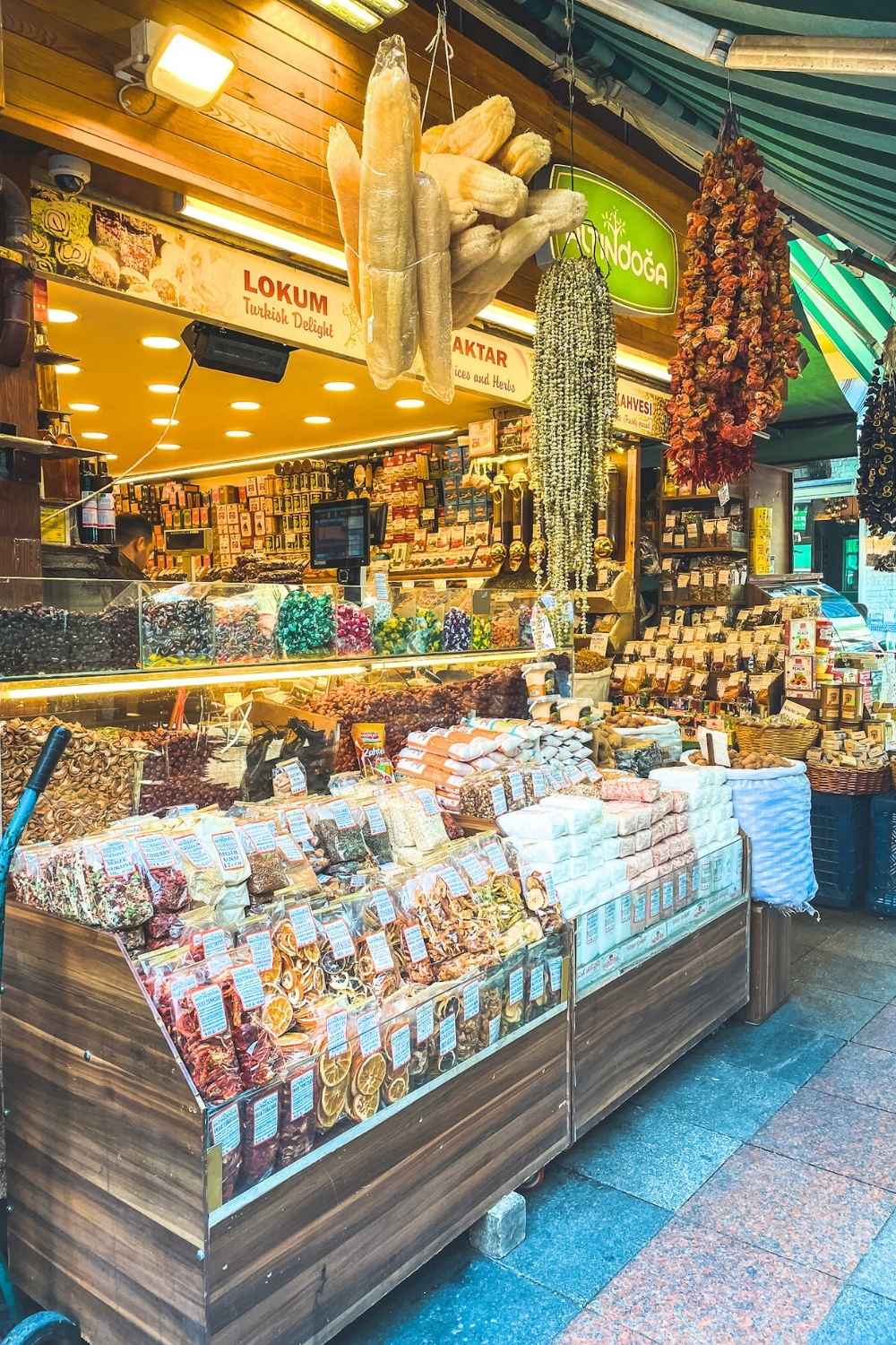 This image features a colorful spice and dried goods stall in Istanbul, showcasing a vibrant assortment of Turkish products. The display includes dried fruits, herbs, spices, teas, and lokum (Turkish delight), with hanging dried peppers and garlic adding a rustic touch. The warm lighting and rich variety of goods highlight the bustling and aromatic atmosphere of a traditional Turkish market.