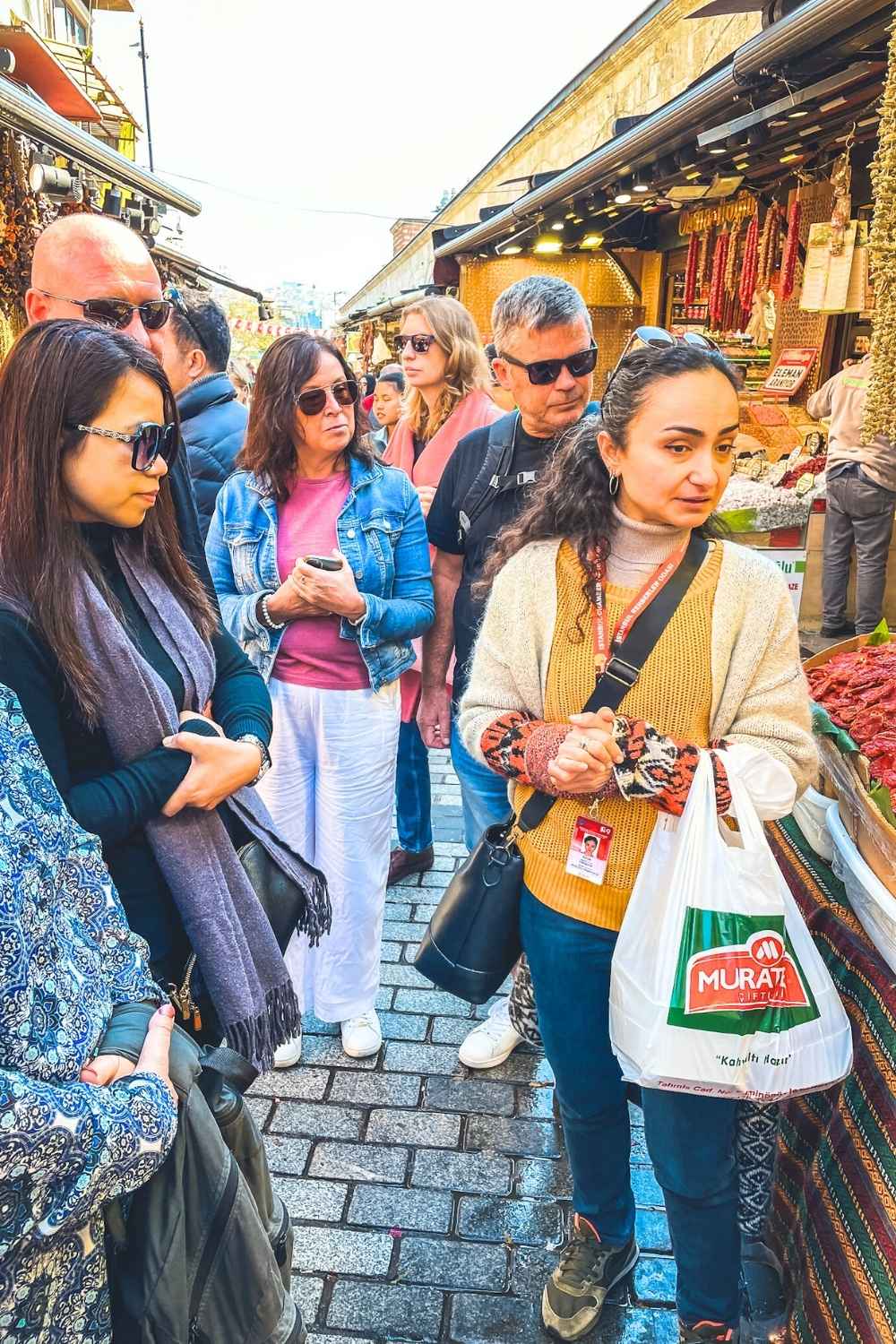 This image shows a group of people gathered at an outdoor market in Istanbul, engaging in a food tour. The guide, holding a shopping bag labeled "Murat," appears to be explaining something, while the group observes stalls filled with dried goods and spices. The bustling atmosphere and vibrant market setting highlight the city’s rich culinary traditions.