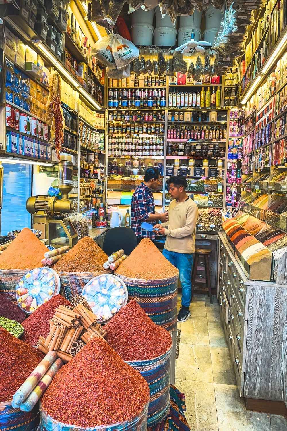 This image showcases a vibrant spice shop in Istanbul, with large barrels overflowing with colorful spices like paprika, turmeric, and cinnamon sticks. Shelves are packed with jars, bottles, and packaged goods, creating a visually rich and aromatic environment. Two shopkeepers are engaged in conversation, emphasizing the authentic and bustling atmosphere of a traditional Turkish bazaar.