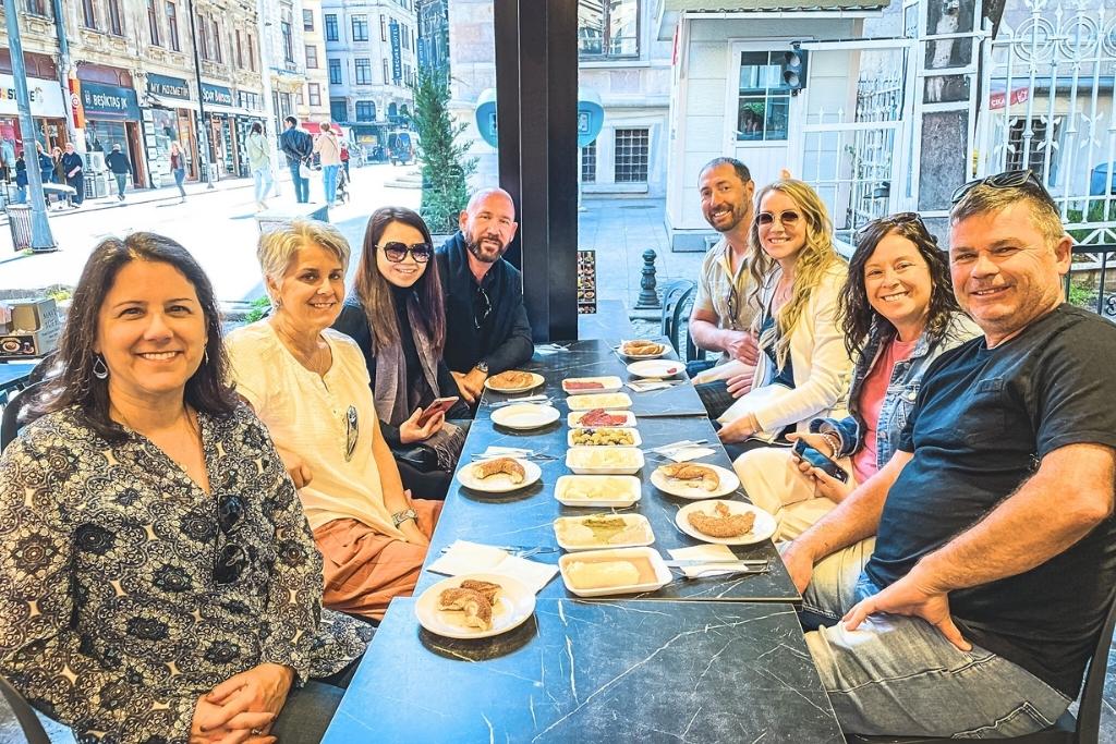 This image features a group of people sitting at a table in a street-side café, enjoying a spread of traditional Turkish dishes during a food tour in Istanbul. The table is set with small plates of assorted foods, and the background shows a charming urban street scene with historic architecture.