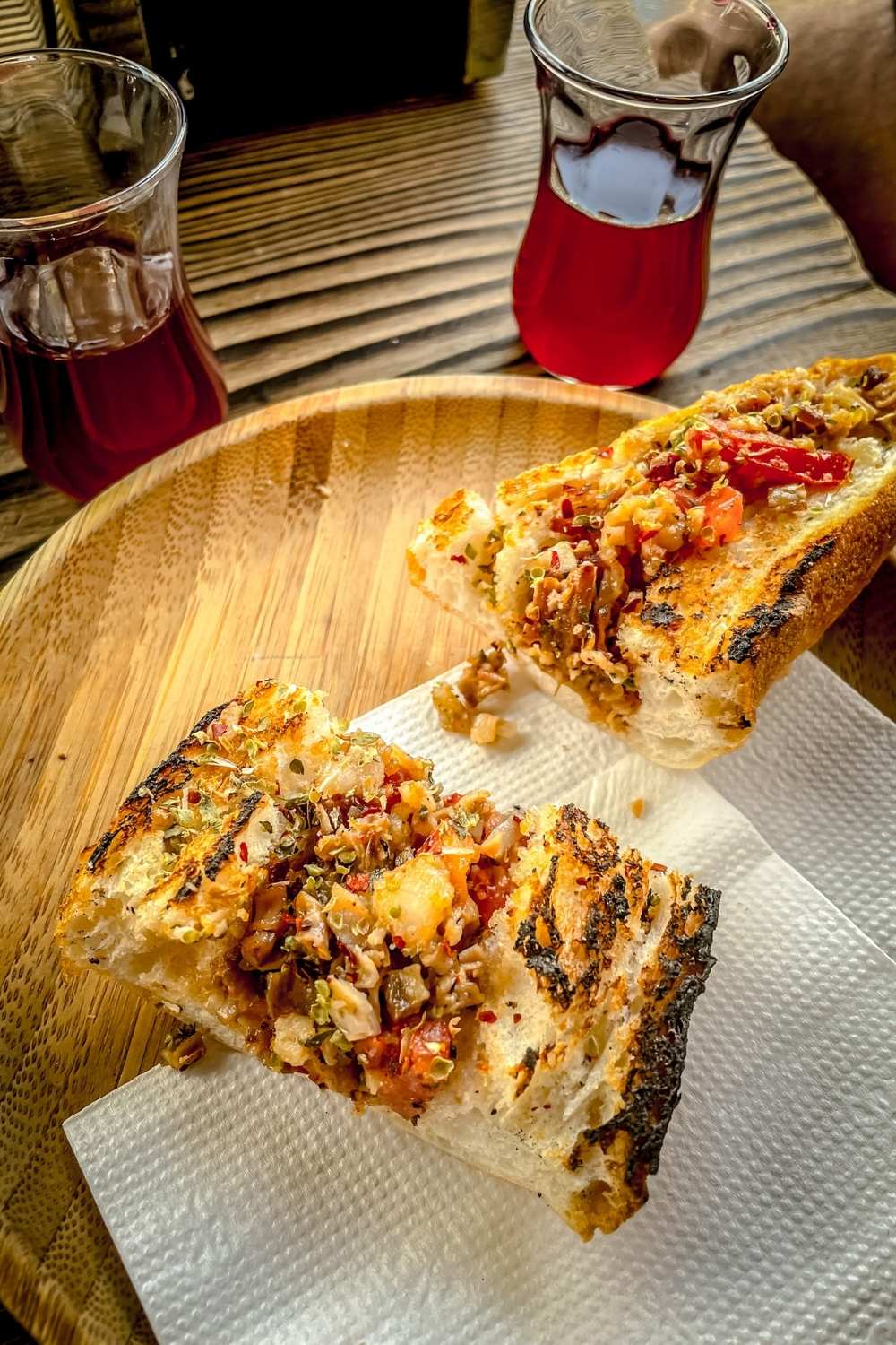 This image highlights two slices of toasted bread topped with a savory mix of tomatoes, herbs, and spices, served on a wooden plate. Accompanying the dish are two glasses of Turkish tea, creating a perfect pairing for a flavorful snack.