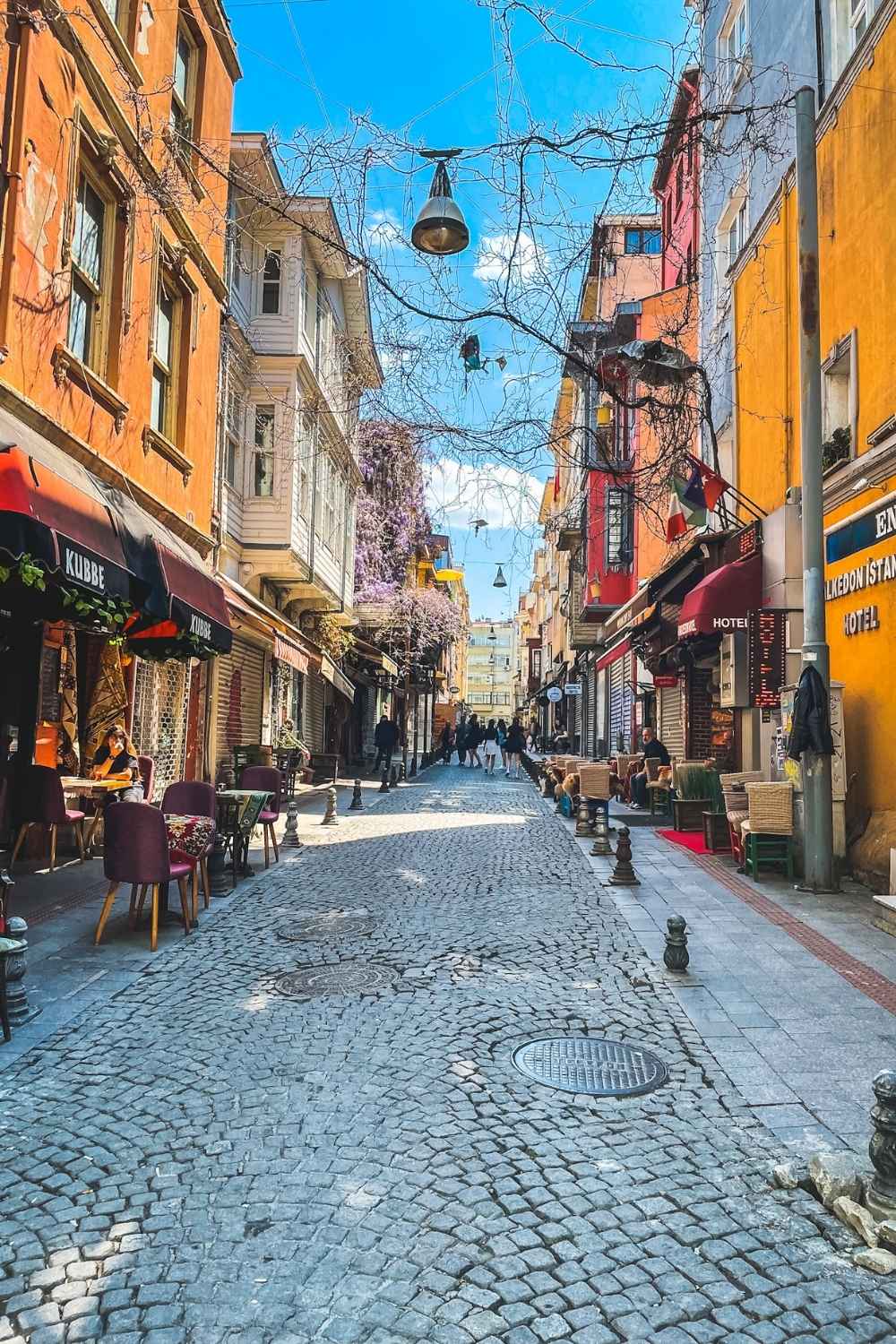 This image captures a picturesque cobblestone street in Istanbul, lined with colorful buildings, quaint cafés, and small shops. The scene exudes charm with vibrant awnings, outdoor seating, and blooming flowers, while people stroll leisurely under a bright blue sky.