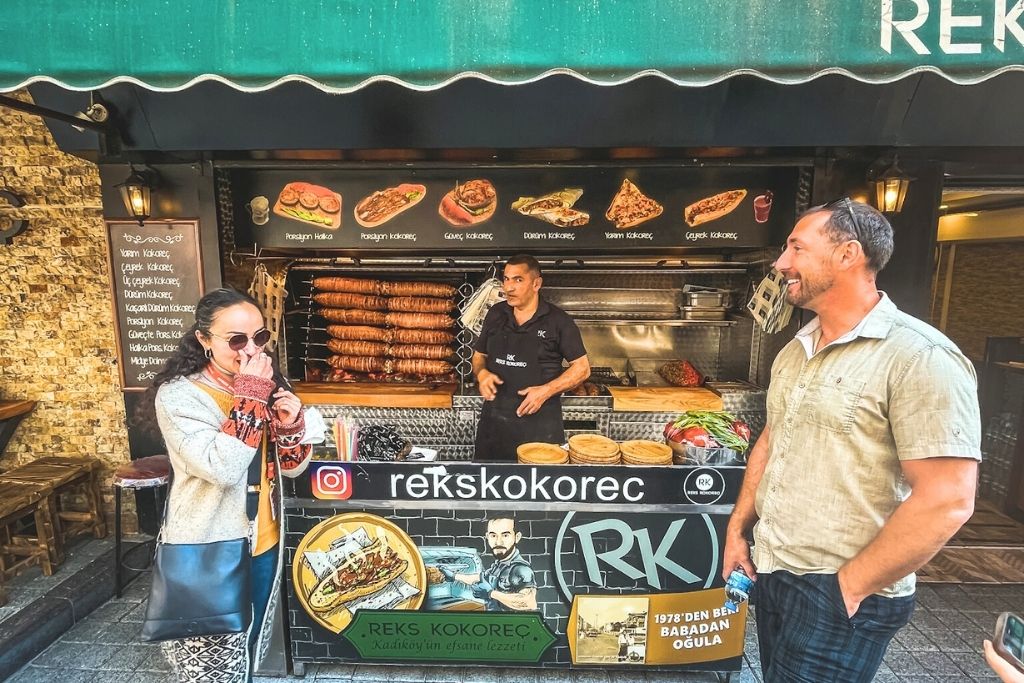 This image shows a vibrant street food stand in Istanbul labeled "Reks Kokorec," specializing in kokoreç, a traditional Turkish dish. The stall displays skewers of grilled meats alongside menu boards featuring various food options, while two people stand in front—one smiling and another enjoying the atmosphere.