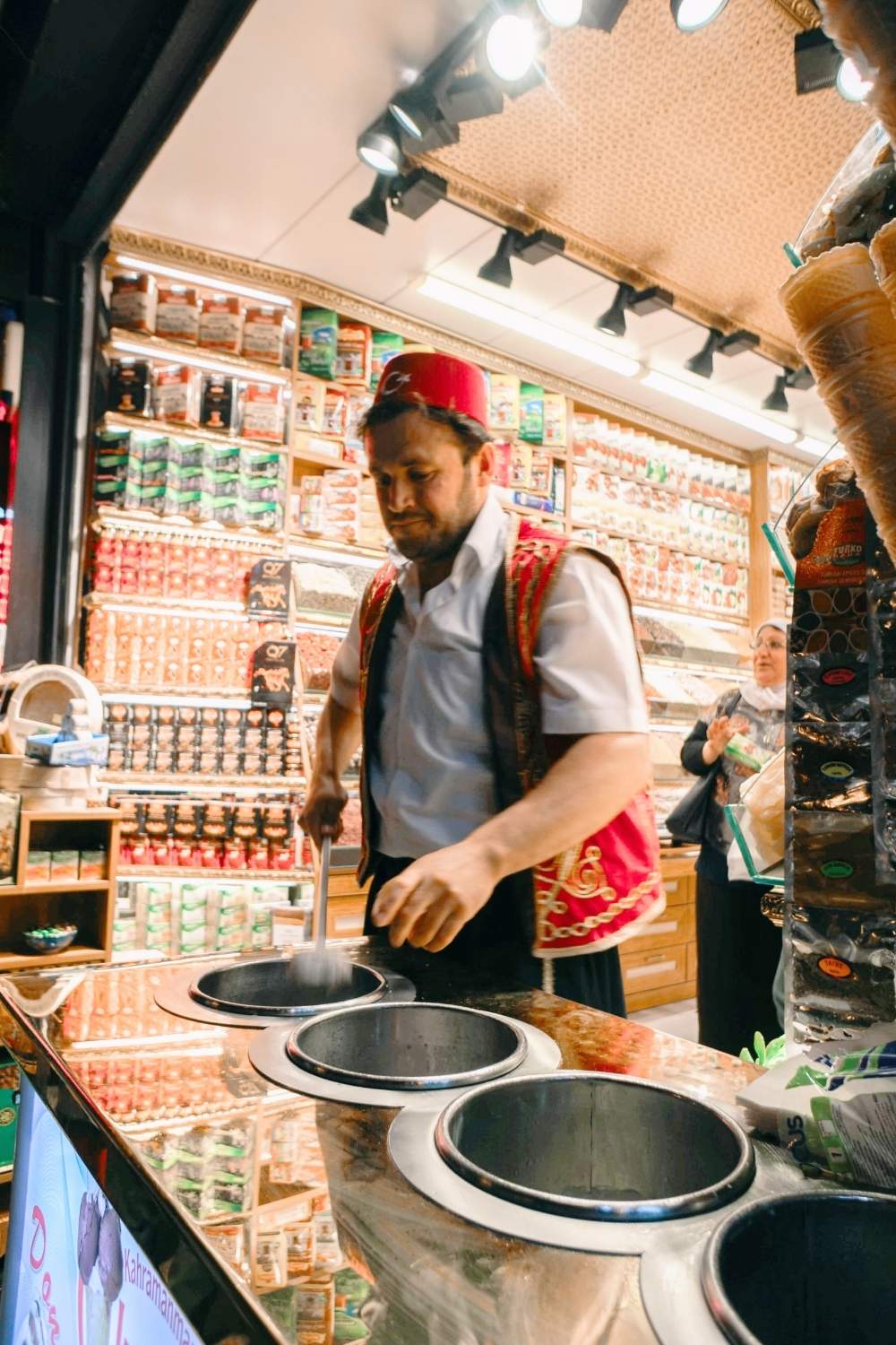 The image shows a Turkish ice cream vendor wearing traditional attire, including a red fez and an embroidered vest, skillfully serving stretchy "dondurma" at a bustling stall.