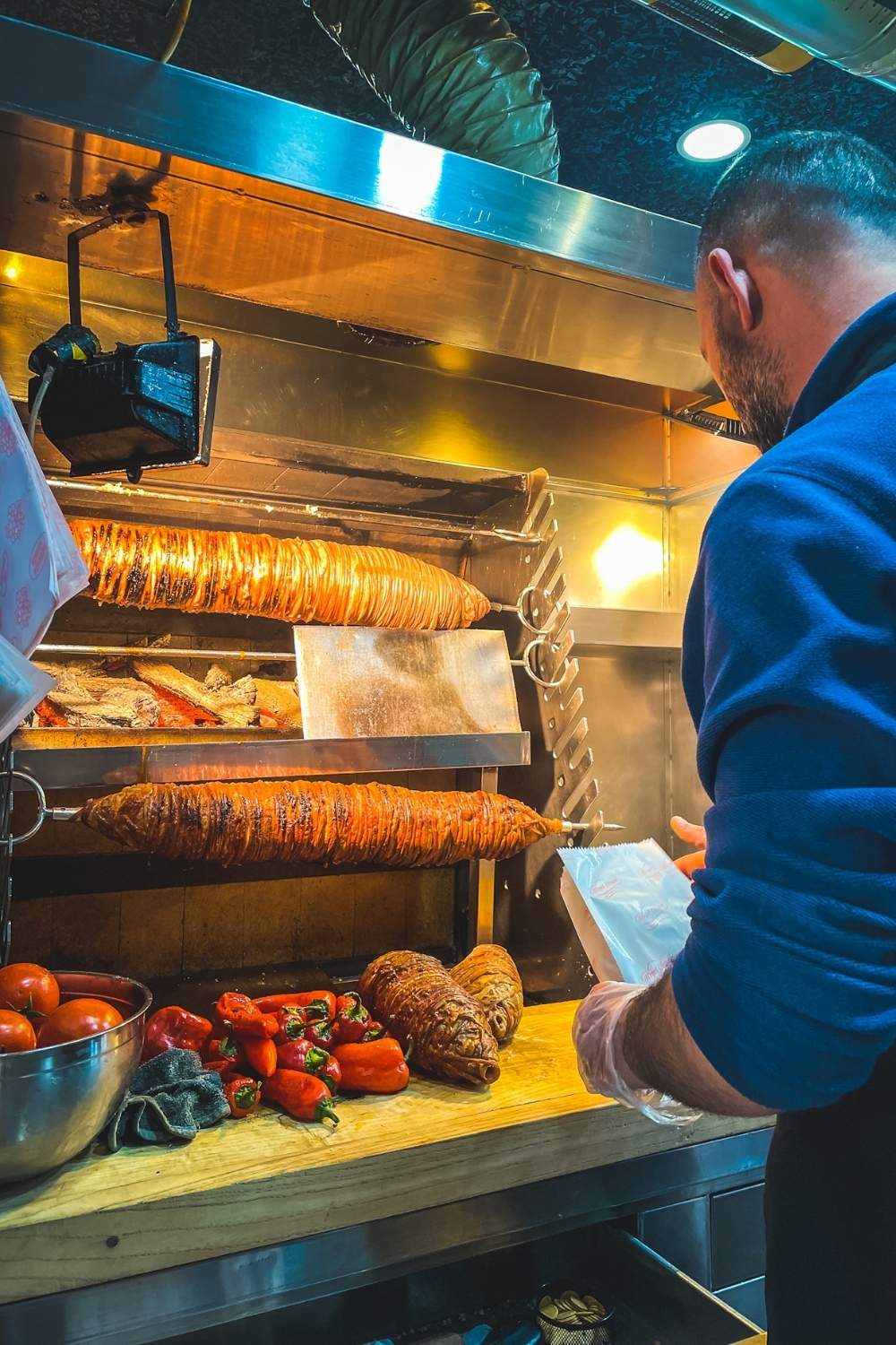 The image shows a vendor preparing kokoreç, a traditional Turkish street food made from spiced and grilled lamb intestines, rotating on a spit. Fresh tomatoes and peppers sit on the counter.