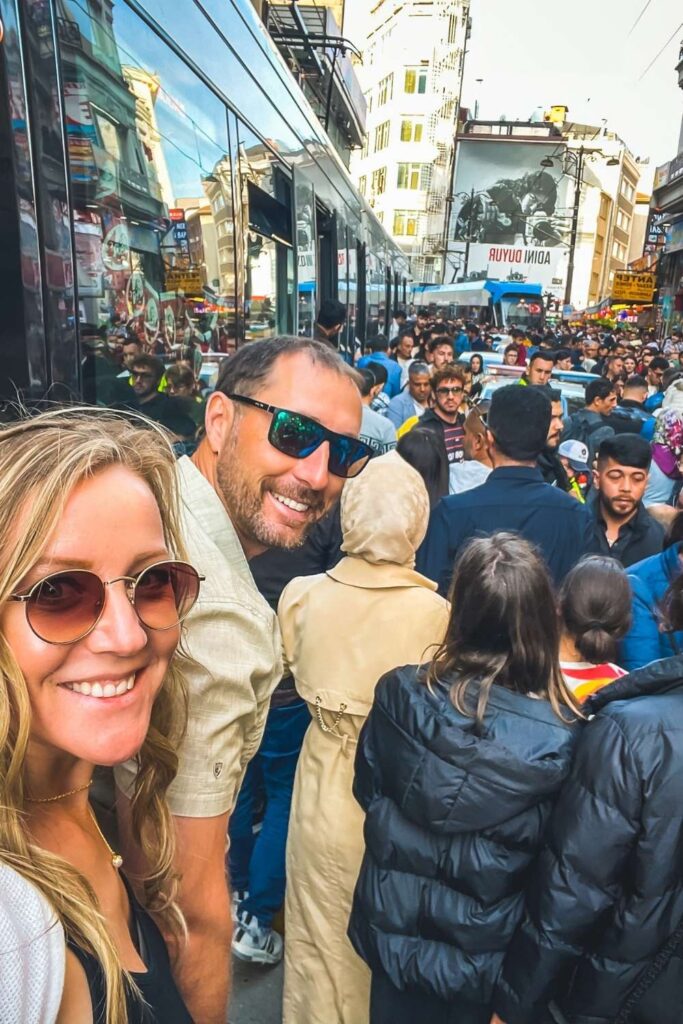 This image shows a crowded street scene in Istanbul with a cheerful Kate and her husband in the foreground taking a selfie. Behind them, a bustling crowd lines the street near a train tram, reflecting the city's lively atmosphere. The mix of modern storefronts and urban architecture in the background completes the energetic vibe of the area.