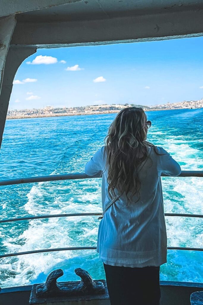 This image captures a serene moment of Kate standing at the back of a ferry, gazing out at the blue waters and distant shoreline. The vibrant sky and gentle waves create a peaceful, scenic backdrop, highlighting the beauty of a ferry ride in Istanbul.