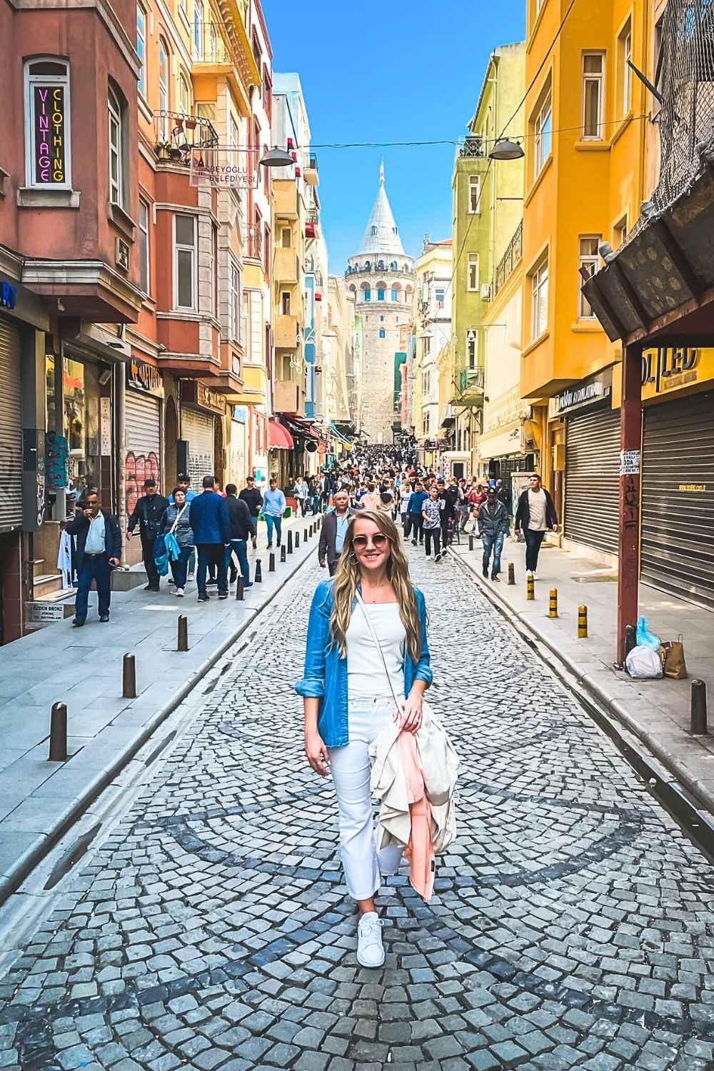 This street scene showcases a cheerful Kate standing on a cobblestone road lined with colorful buildings in Istanbul, with the iconic Galata Tower visible in the background. The lively street is filled with locals and tourists, reflecting the bustling atmosphere of this historic area. The bright colors and clear blue sky create an inviting urban setting.