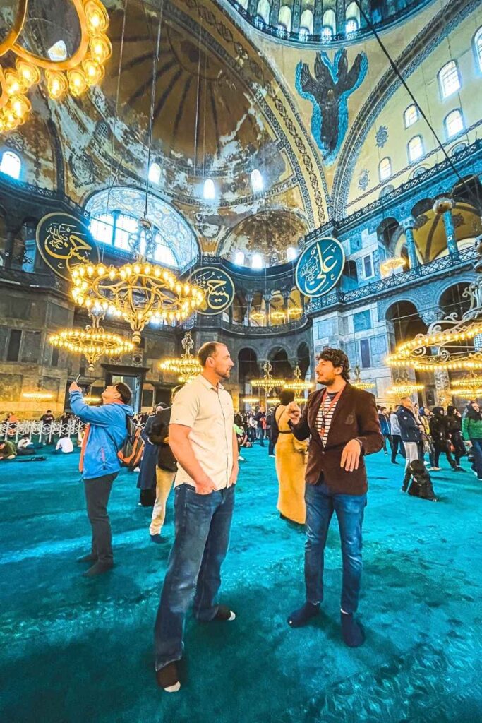 This image captures a vibrant interior scene of the Hagia Sophia in Istanbul, showcasing its grand domes, intricate Islamic calligraphy, and golden chandeliers. In the foreground, two men are engaged in conversation, with one who appears to be a tour guide gesturing animatedly.