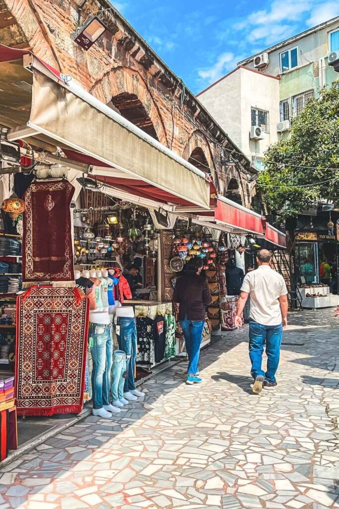 This image captures a charming outdoor market in Istanbul, with colorful stalls showcasing items like rugs, lanterns, and clothing. The cobblestone pathway and historic brick arches in the background add a rustic charm, while visitors stroll and browse the vibrant displays. 