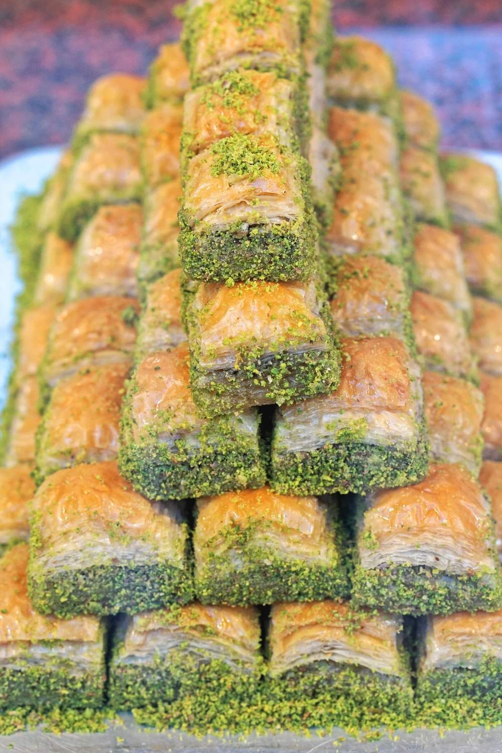 The image displays a pyramid stack of Turkish pistachio baklava, showcasing layers of golden, flaky pastry generously coated with vibrant green ground pistachios.