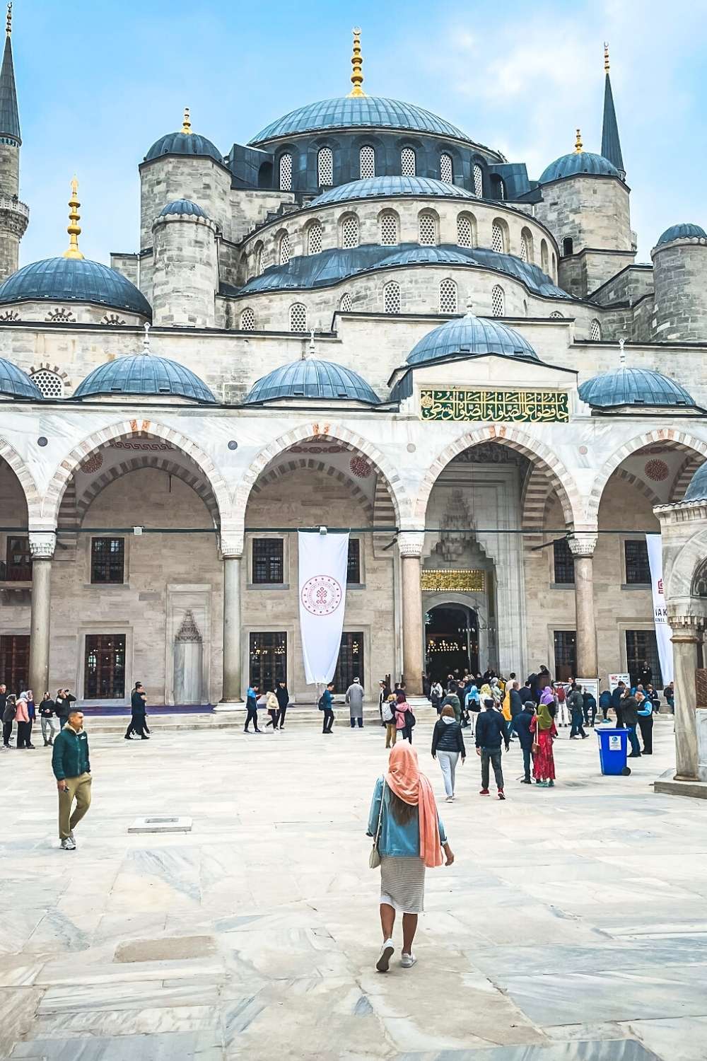 This image showcases the courtyard of the Blue Mosque (Sultan Ahmed Mosque) in Istanbul, with its grand domes, intricate arches, and Islamic calligraphy above the entrance. Visitors wander through the expansive marble courtyard, admiring the historic architecture. Kate, in the foreground, wearing a light pink scarf walks toward the mosque.