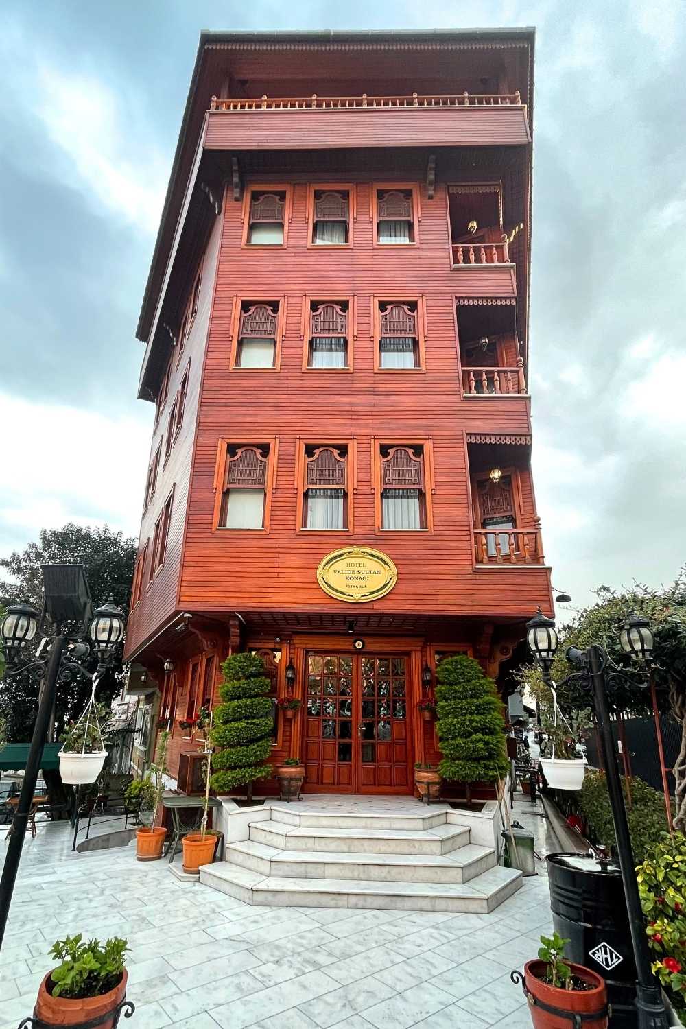 This image shows a charming, traditional-style wooden hotel named "Hotel Valide Sultan Konagi" in Istanbul. The building's warm red facade, adorned with decorative window shutters and balconies, contrasts beautifully with the lush greenery and potted plants at the entrance. The marble steps and ornate lamps add a touch of elegance, set against a cloudy sky that enhances the inviting atmosphere.