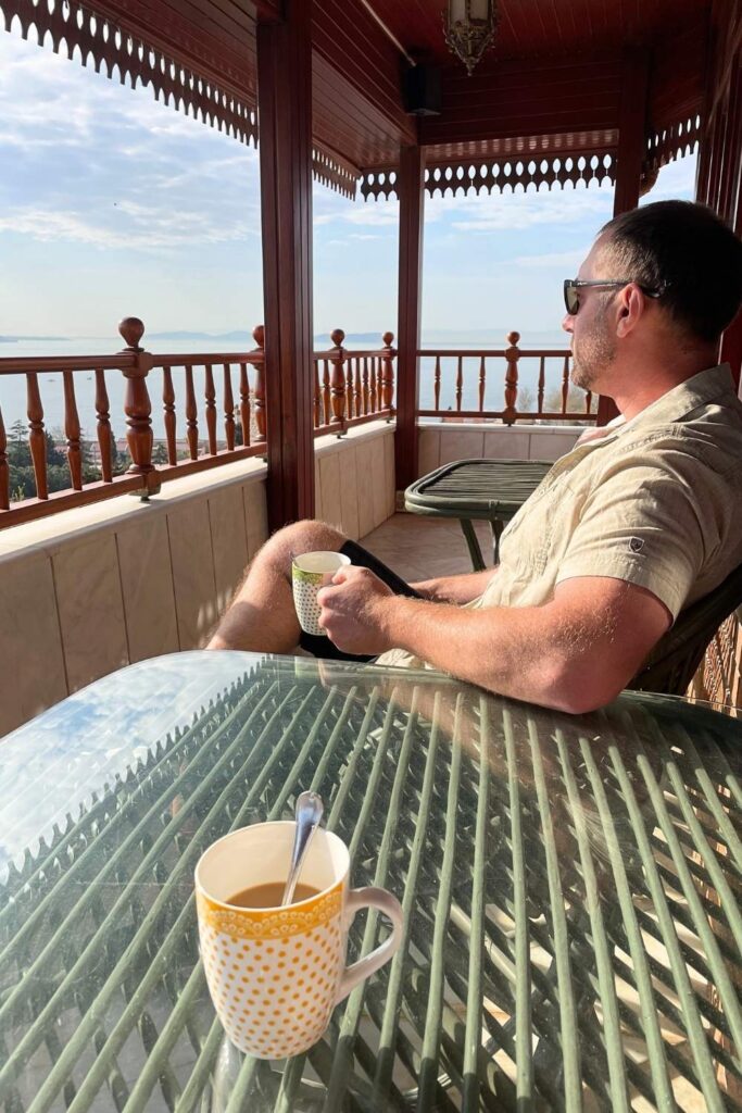 Kate's husband, wearing sunglasses and a beige shirt, relaxes on a wooden balcony with ornate railings, sipping coffee while enjoying a serene sea view.