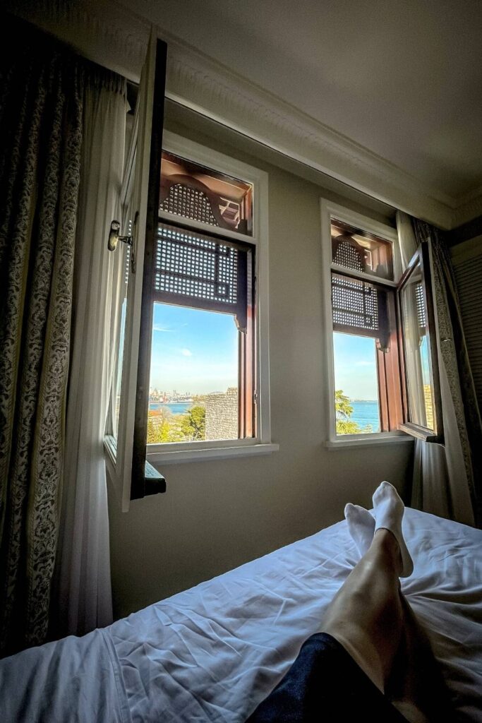 This cozy image captures a relaxed moment, with someone lying on a bed, feet up, looking out through open windows at a stunning view of the sea and distant cityscape. The traditional wooden window frames and light curtains create a warm and inviting interior, while the bright blue sky and ocean outside add a sense of tranquility and escape. The perspective highlights a peaceful retreat with a breathtaking view.