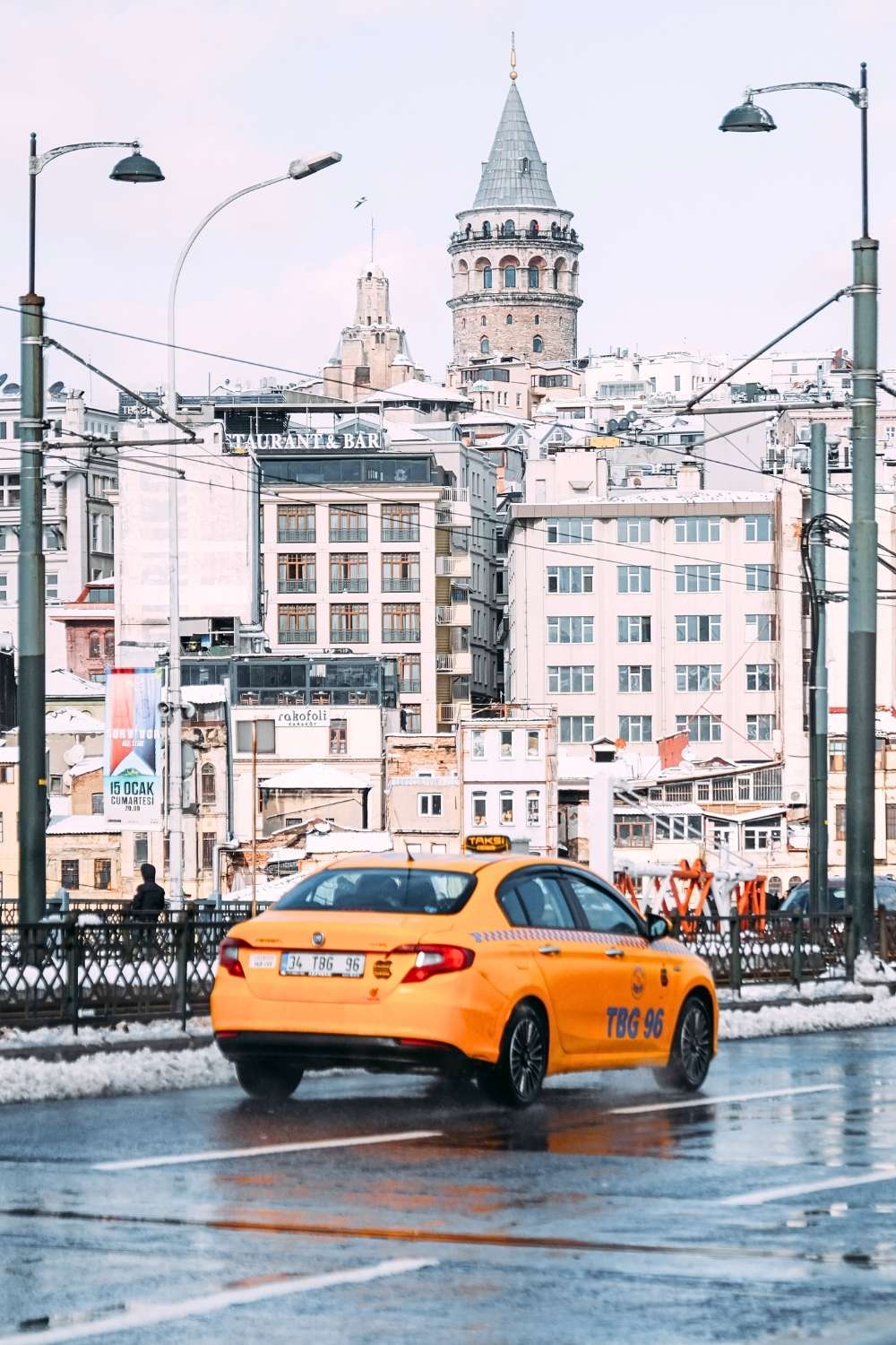 This image showcases a rainy street in Istanbul, with a bright yellow taxi in the foreground reflecting on the wet pavement. In the background, the iconic Galata Tower rises above the cityscape, surrounded by historic and modern buildings.