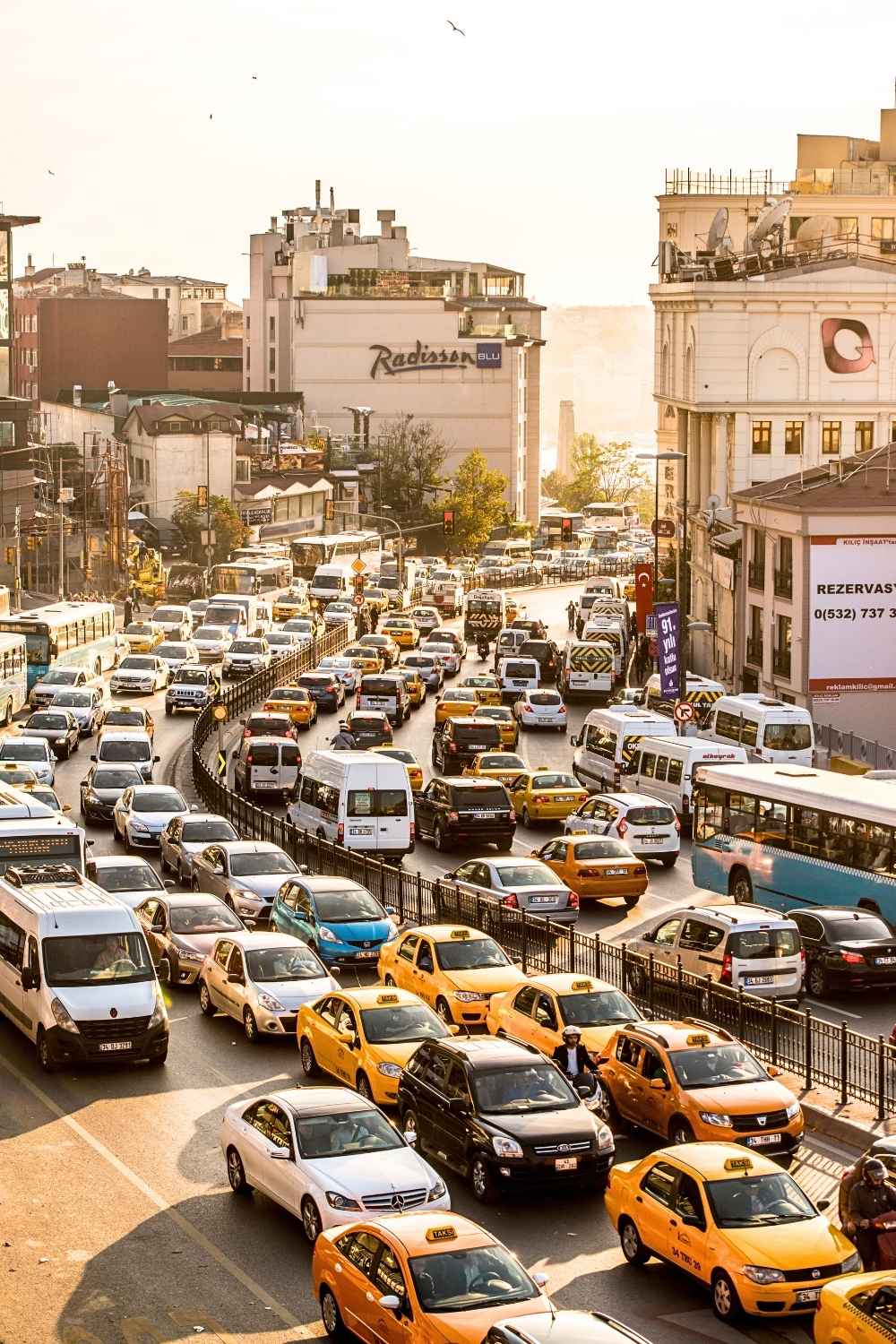 This image captures a busy traffic scene in Istanbul during golden hour, with yellow taxis, buses, and private vehicles crowding the road. The warm sunlight bathes the urban landscape, highlighting nearby buildings, including a visible "Radisson Blu" hotel.